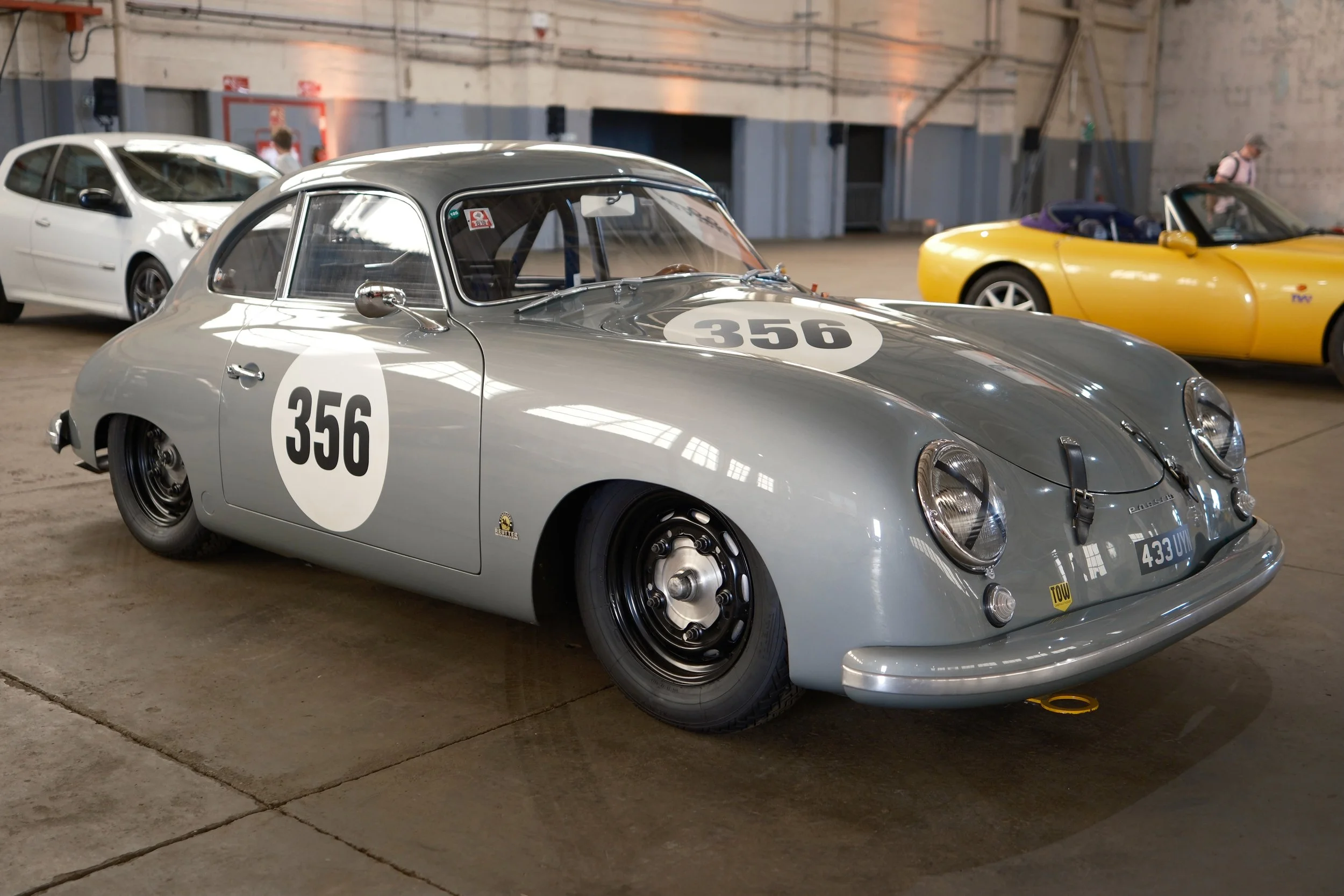 A vintage silver race car with the number 356 parked indoors at a car show, with a white modern car and a yellow sports car in the background.