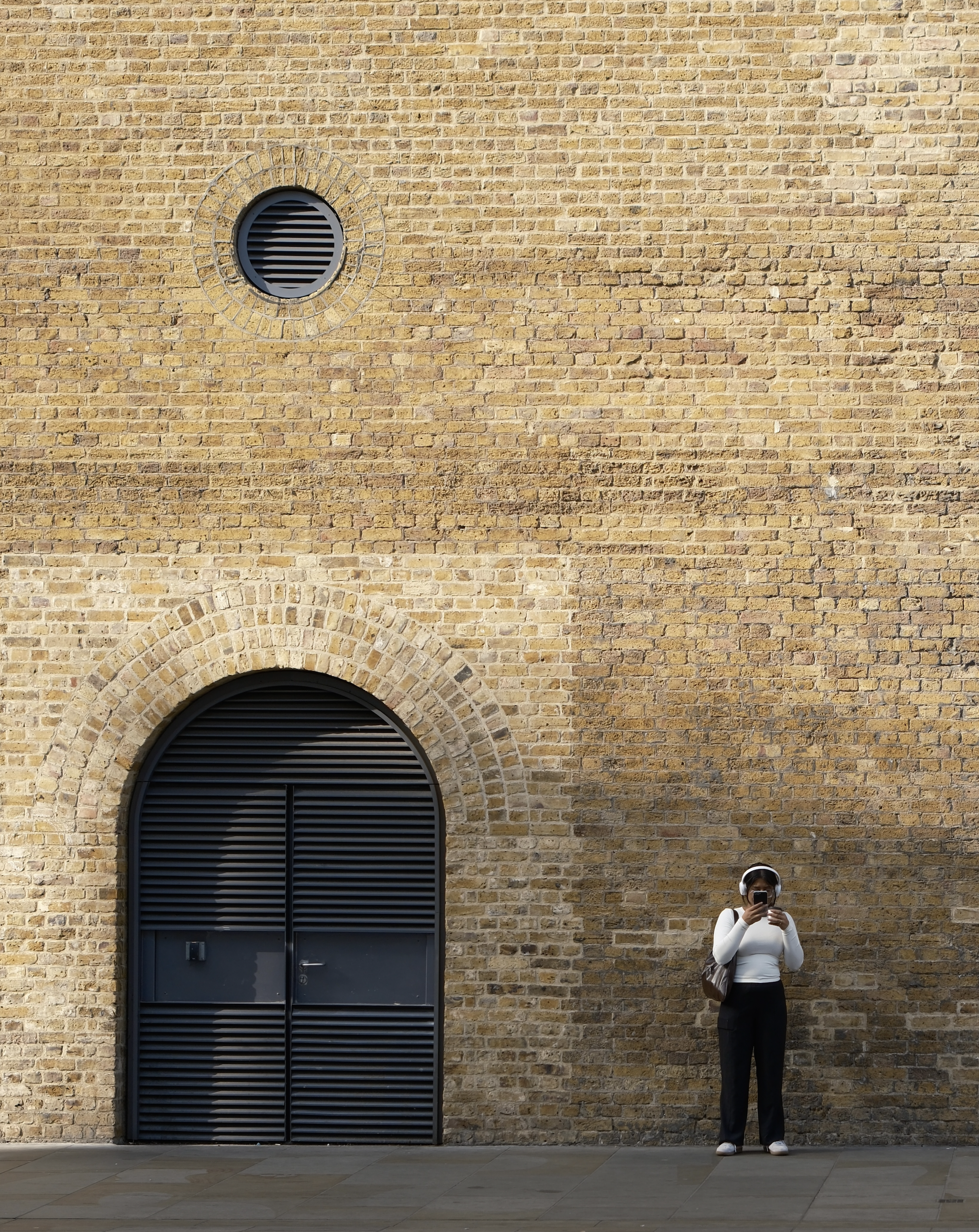 A woman in white top and black pants taking a selfie with her phone, standing in front of a brick wall with a large arched window with black blinds and a small round vent above it.