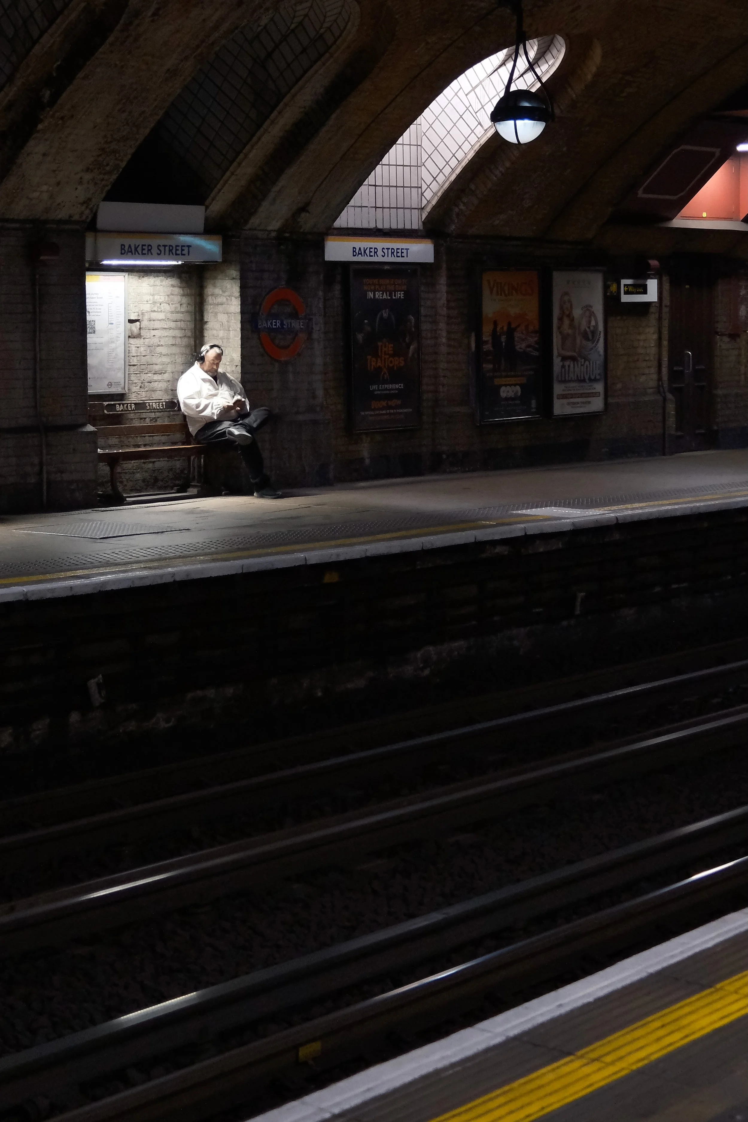 A man sitting on a bench at a subway station, wearing headphones and using his mobile phone. The station has brick walls, signs reading 'Baker Street', and movie posters on the wall. The platform has yellow safety lines and tracks below.