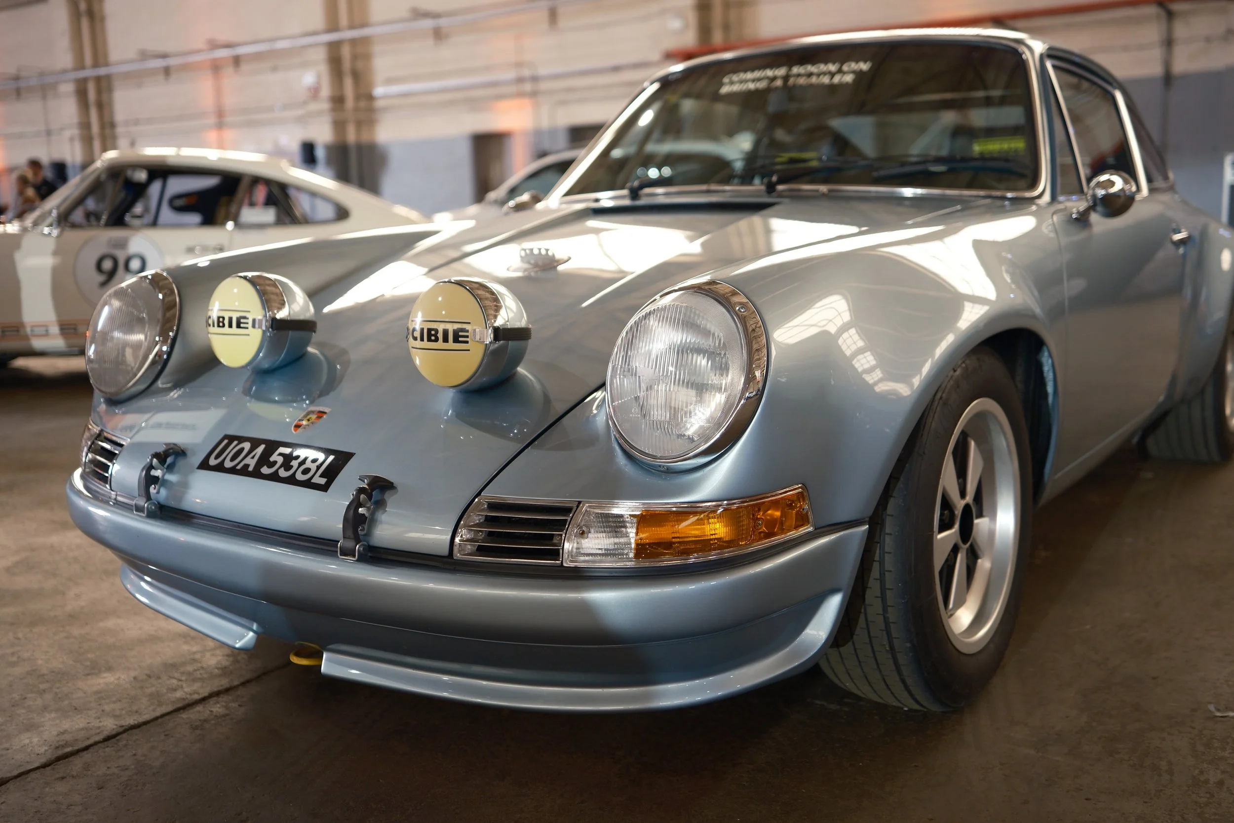 A vintage light blue Porsche 911 sports car with rally lights on the front, parked indoors at a car exhibition.