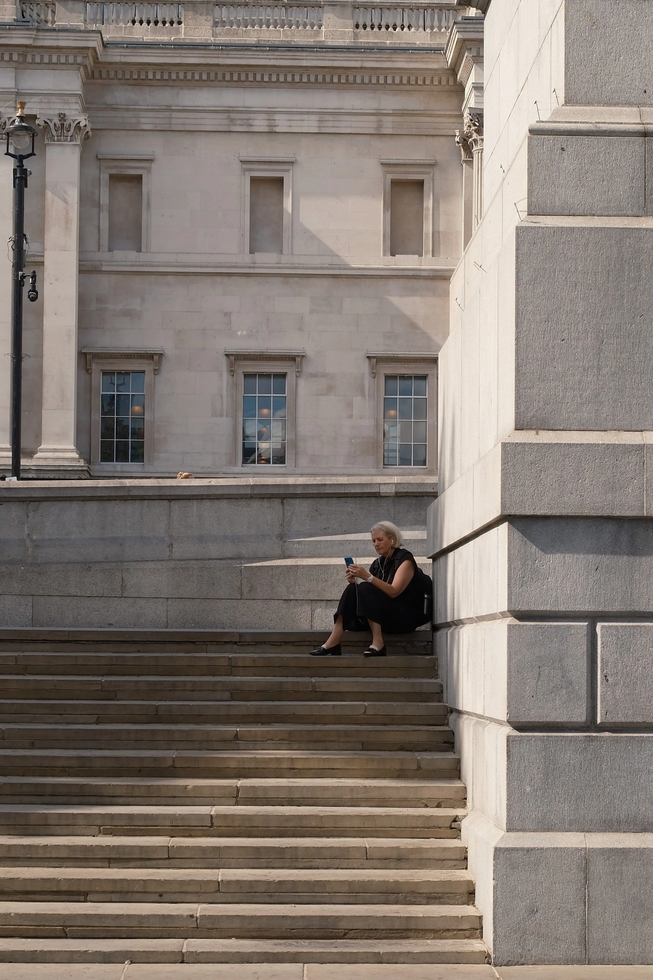 A woman seated on stone steps outside a historic building, looking at her phone, with an ornate façade featuring windows and columns in the background.