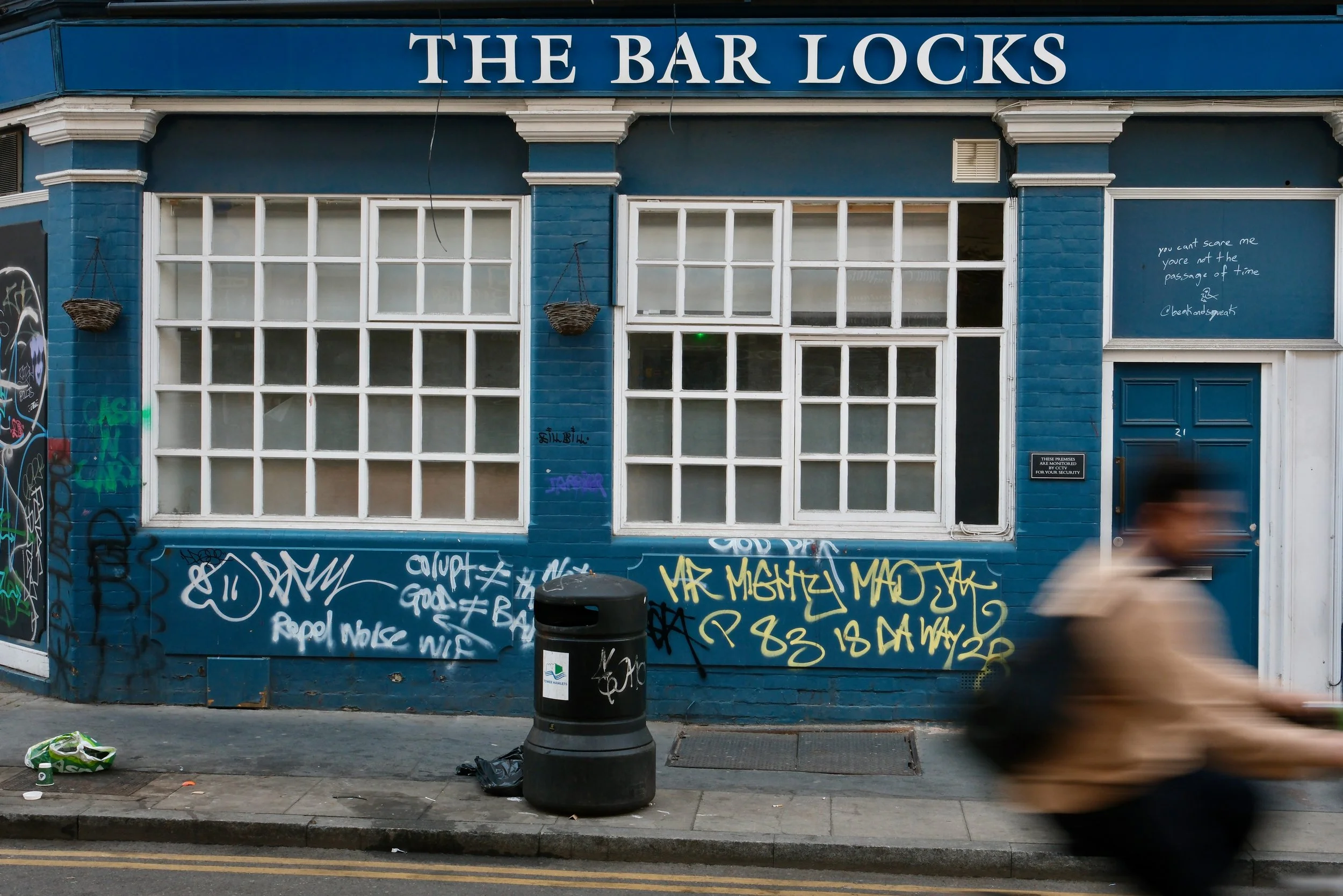 Blue storefront building with large sign reading 'THE BAR LOCKS' and white window frames. Graffiti on the lower part of the wall and a trash bin in front. A person walking past in motion blur.