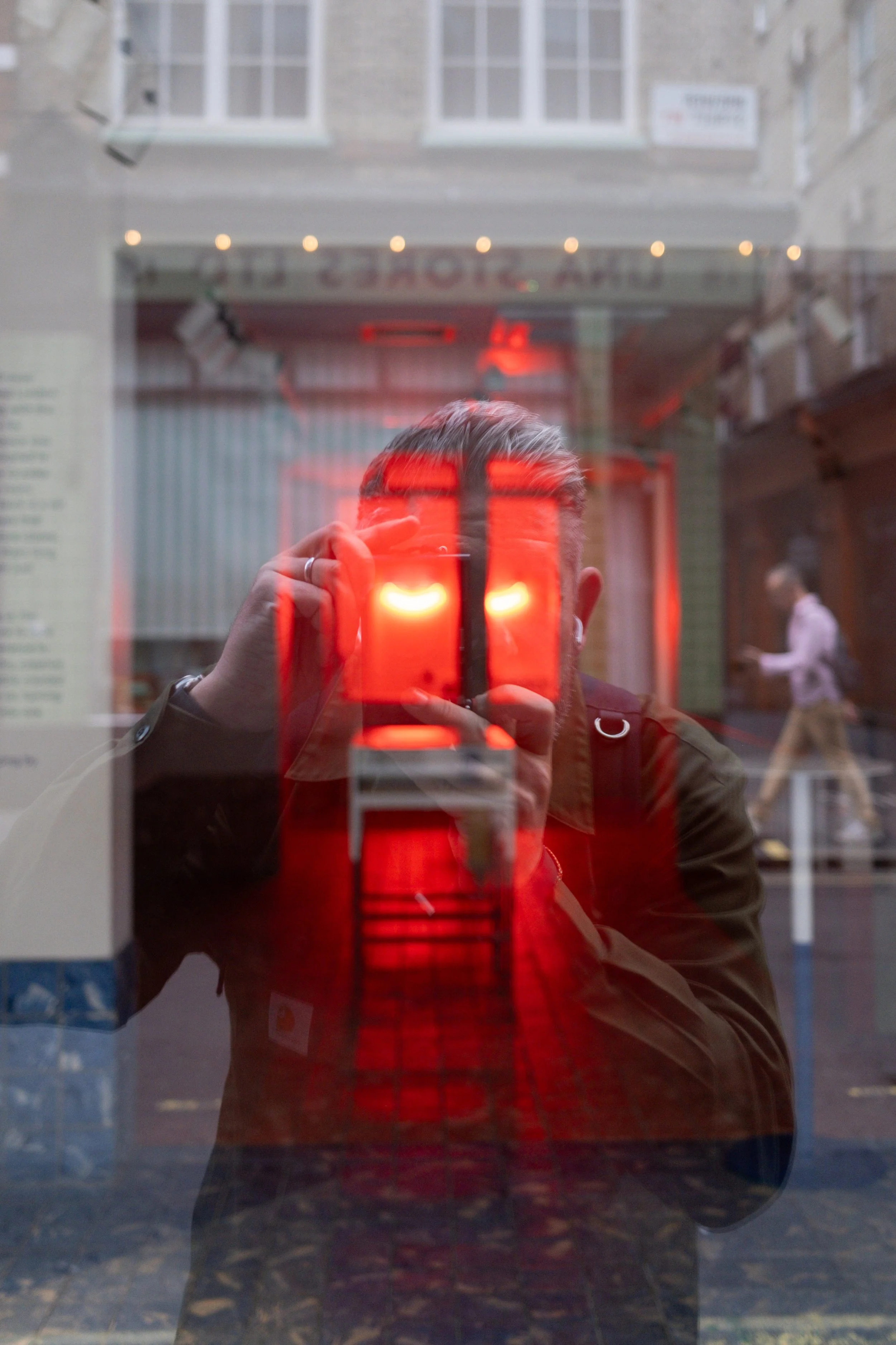Reflection of a person holding a phone, taking a photo through a glass window with a red illuminated face image displayed on the screen. The person has short hair, earrings, and is dressed in a jacket. The background shows a city street with a pedest