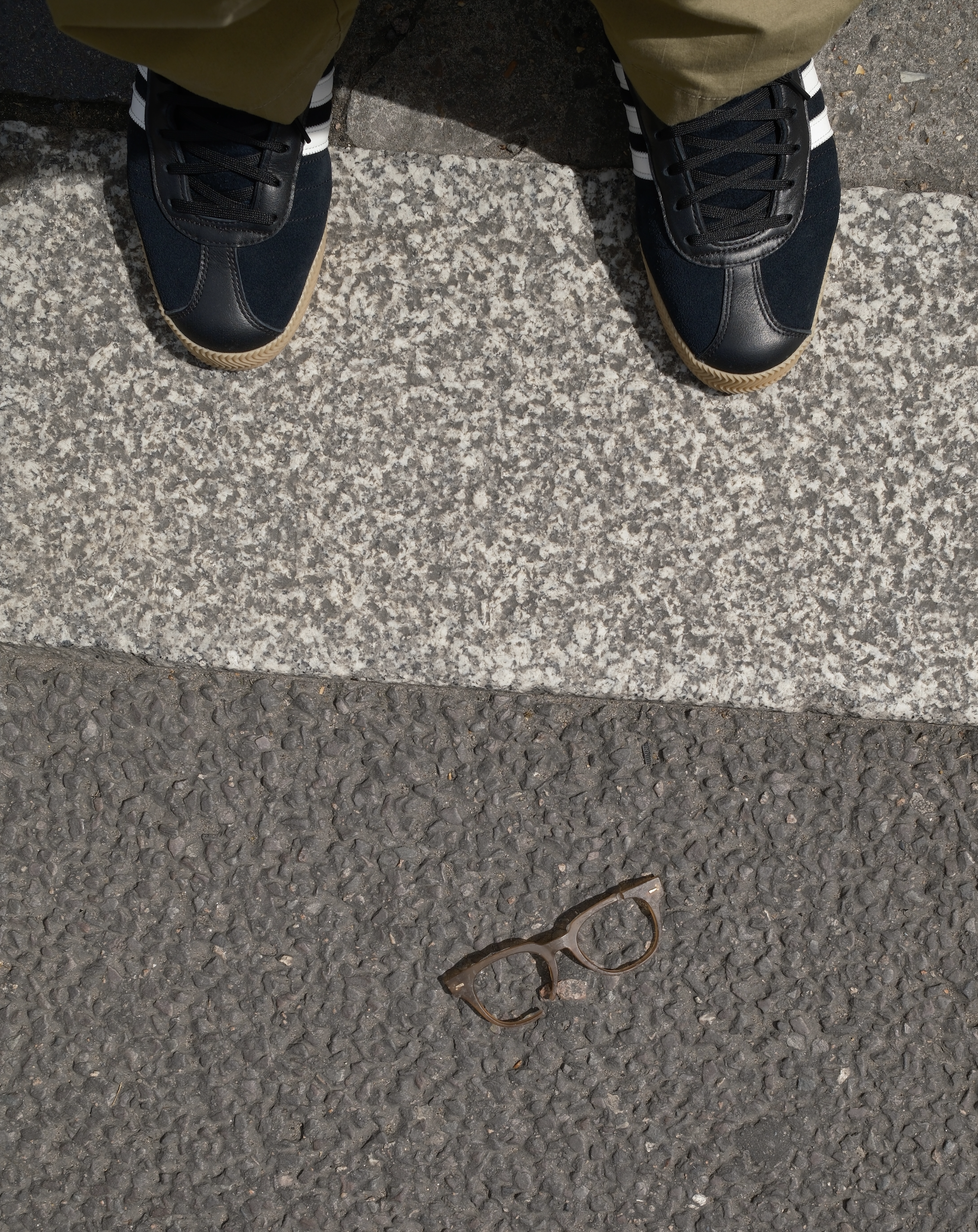 Looking down at a pair of black and navy suede sneakers with gum soles, worn with olive green pants. The shoes are on a curb next to a gray textured sidewalk with a pair of brown tortoise shell glasses on the ground.