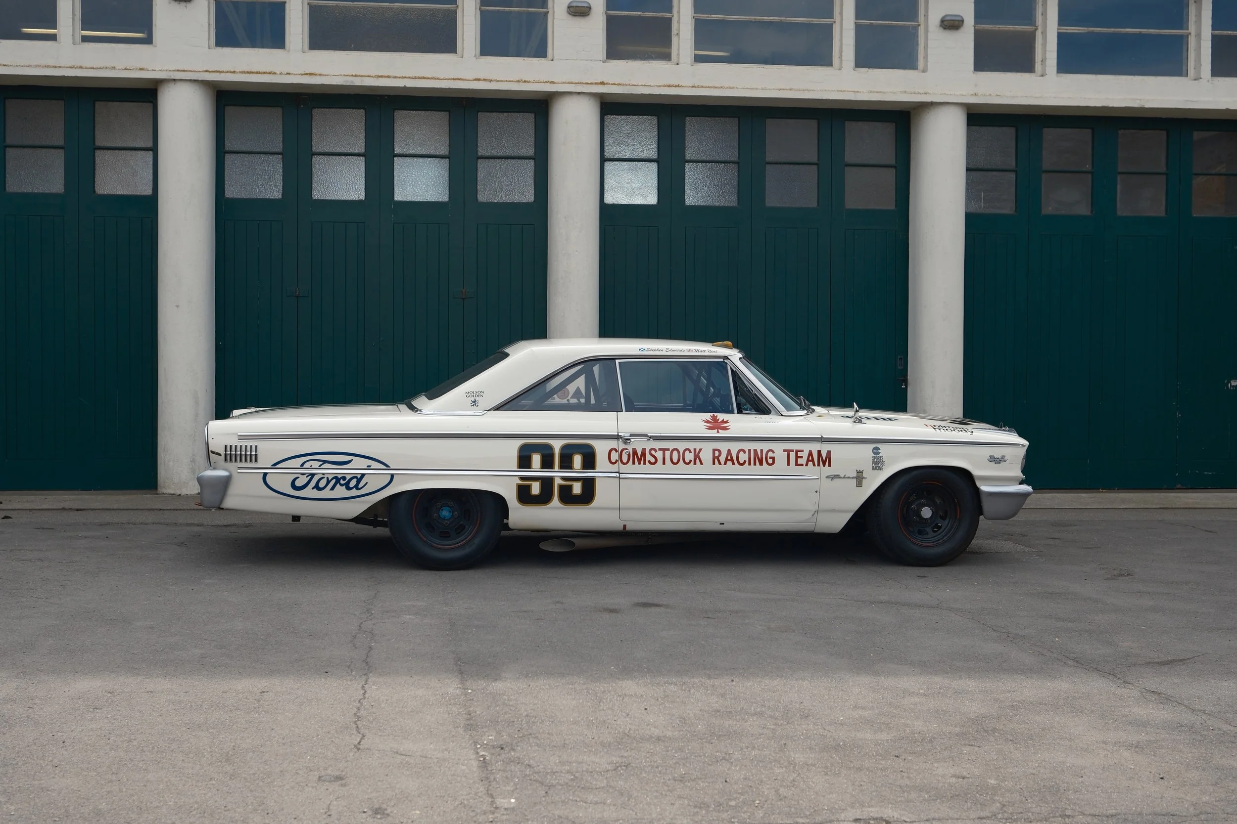 A vintage white race car with the number 99 and 'Comstock Racing Team' written on the side, parked in front of a green garage door with large white columns and upper windows.