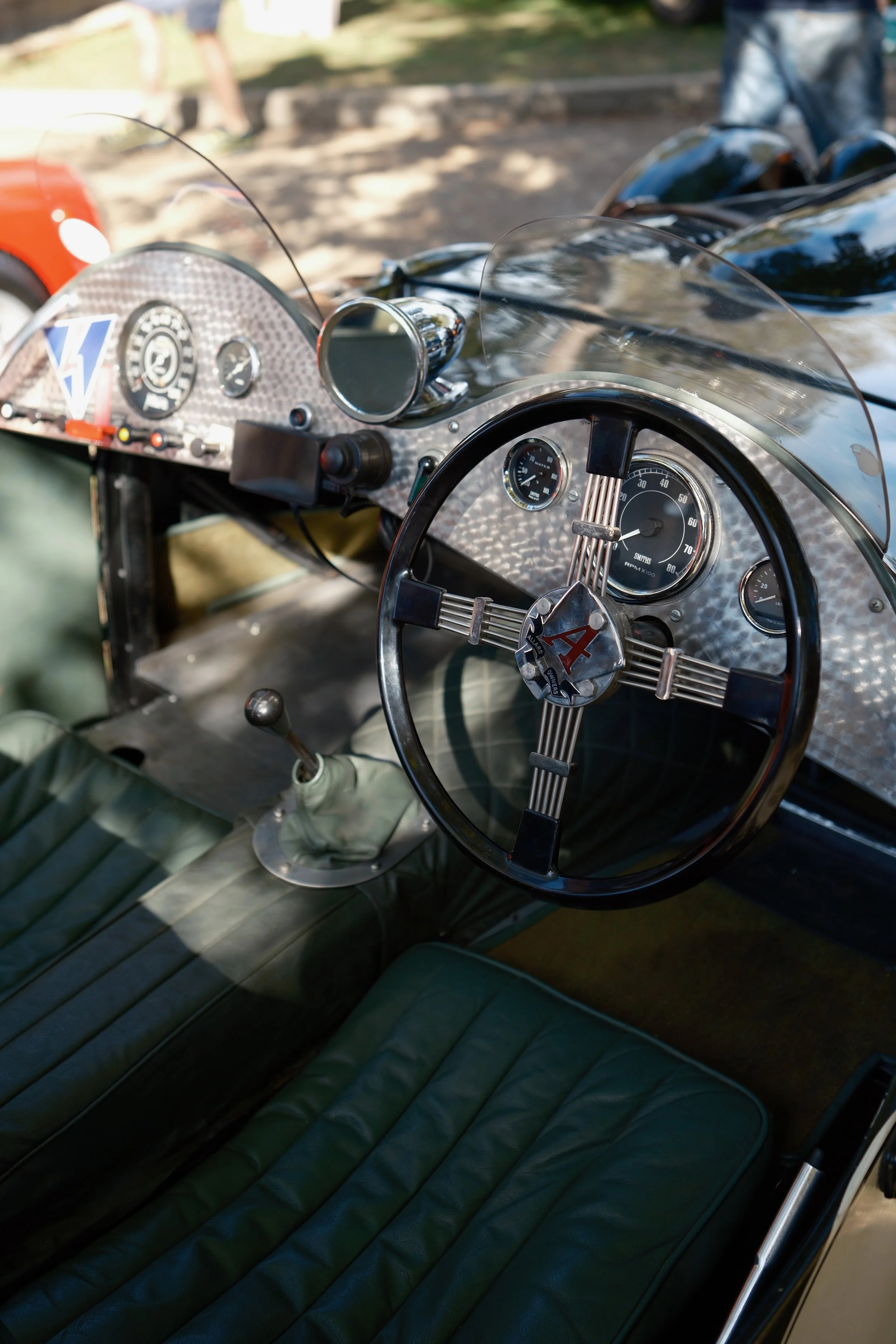 Close-up of a vintage race car interior, showing a wooden steering wheel with an 'Aston Martin' logo in the center, a speedometer, tachometer, and vintage dashboard gauges, with green leather seats.