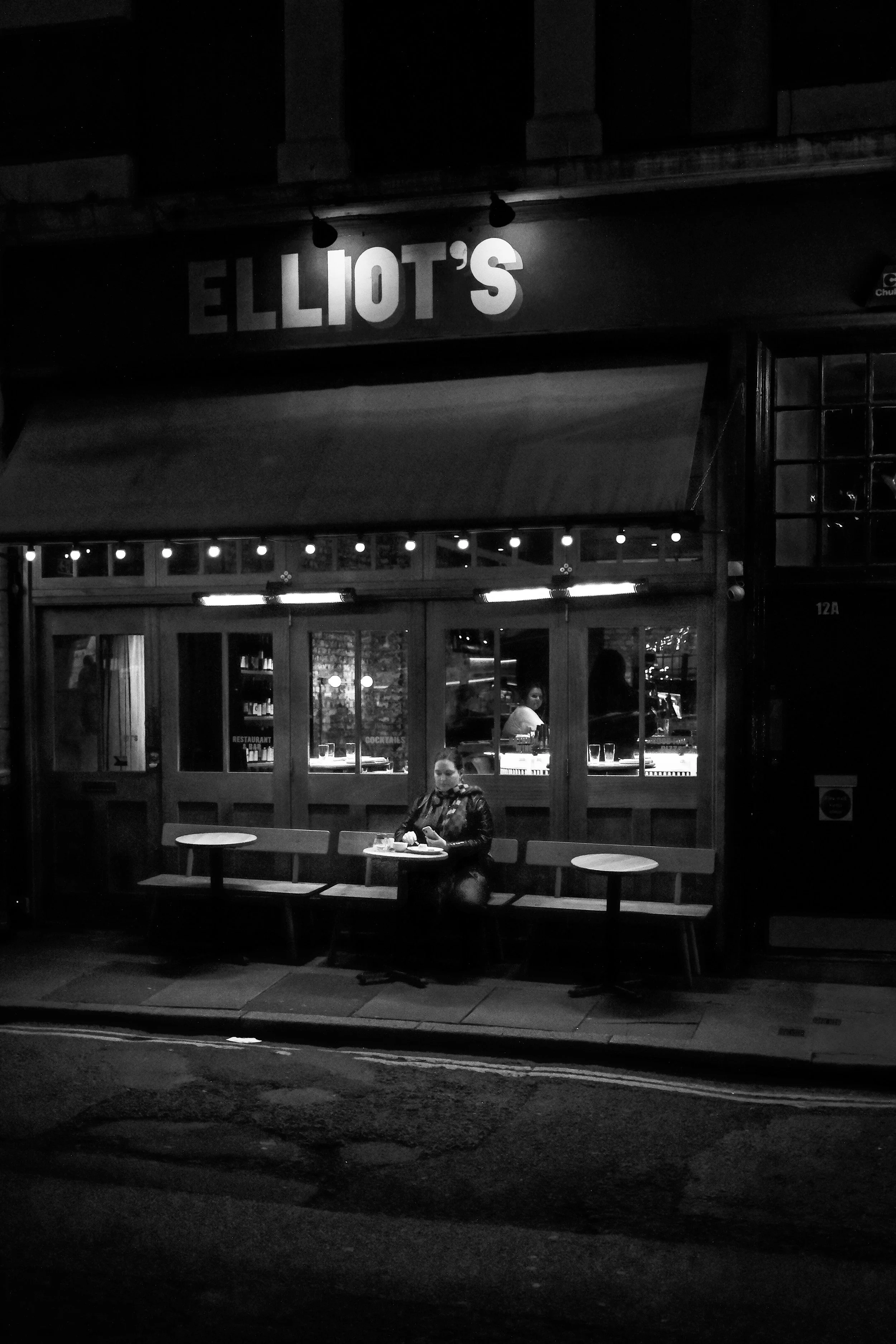 A black and white photo of a woman sitting alone at a table outside a restaurant named Elliot's. The restaurant's interior can be seen through large windows, with a bartender or server visible inside. The woman is looking at her phone and there are e