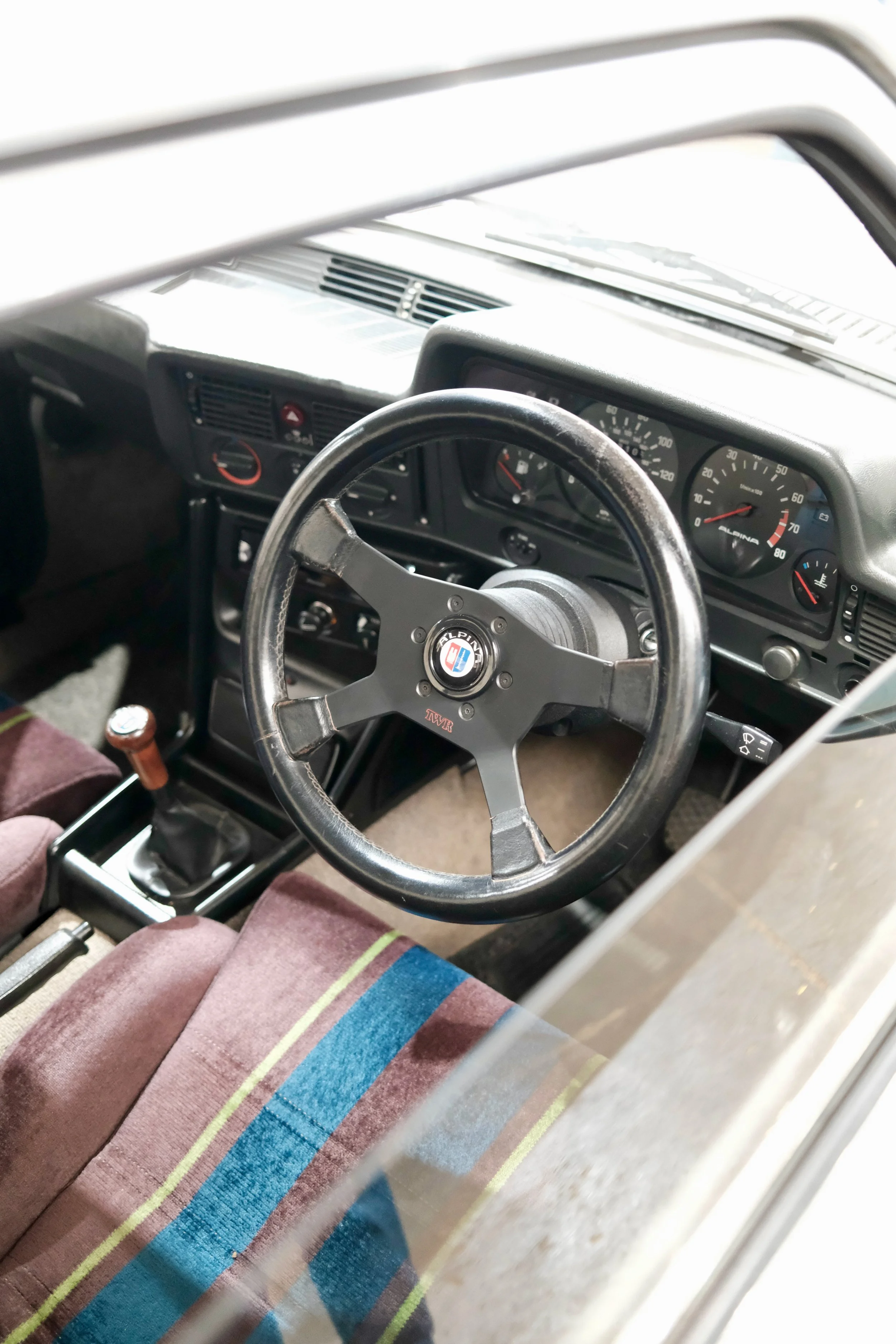 View of the interior of a vintage car, including the steering wheel, dashboard, and gear shift. The steering wheel has an Alpine badge, and the dashboard features analog gauges.