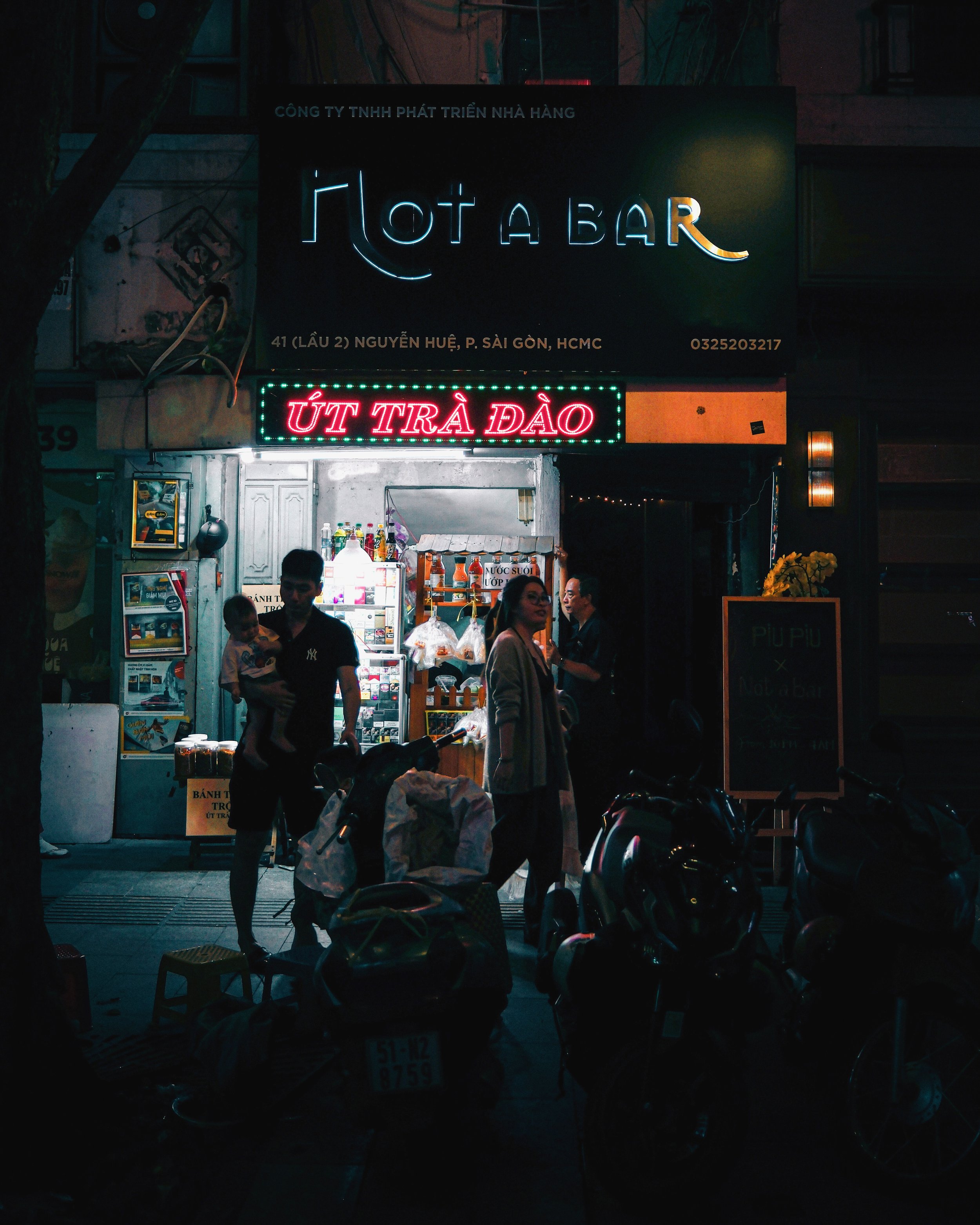Night scene of an outdoor street food stall with neon signs in Vietnamese. The upper black sign displays white text, while a red neon sign below advertises strawberry milk. There are people standing near the stall, and parked motorbikes in front of i