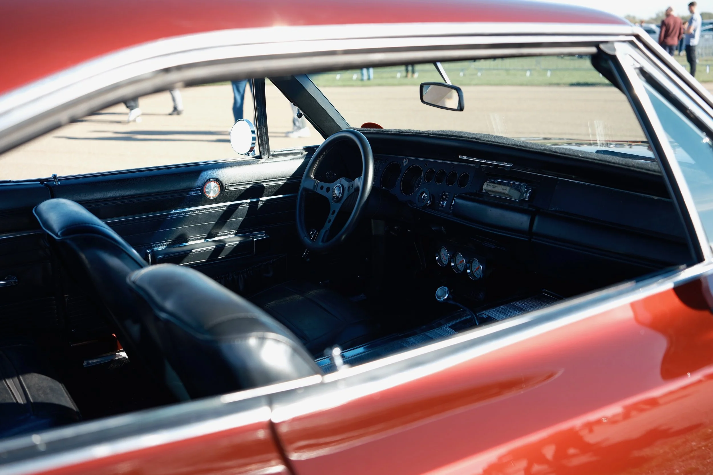 Interior view of a vintage red car showing the dashboard, steering wheel, and front seats, with a few people standing outside in the background.