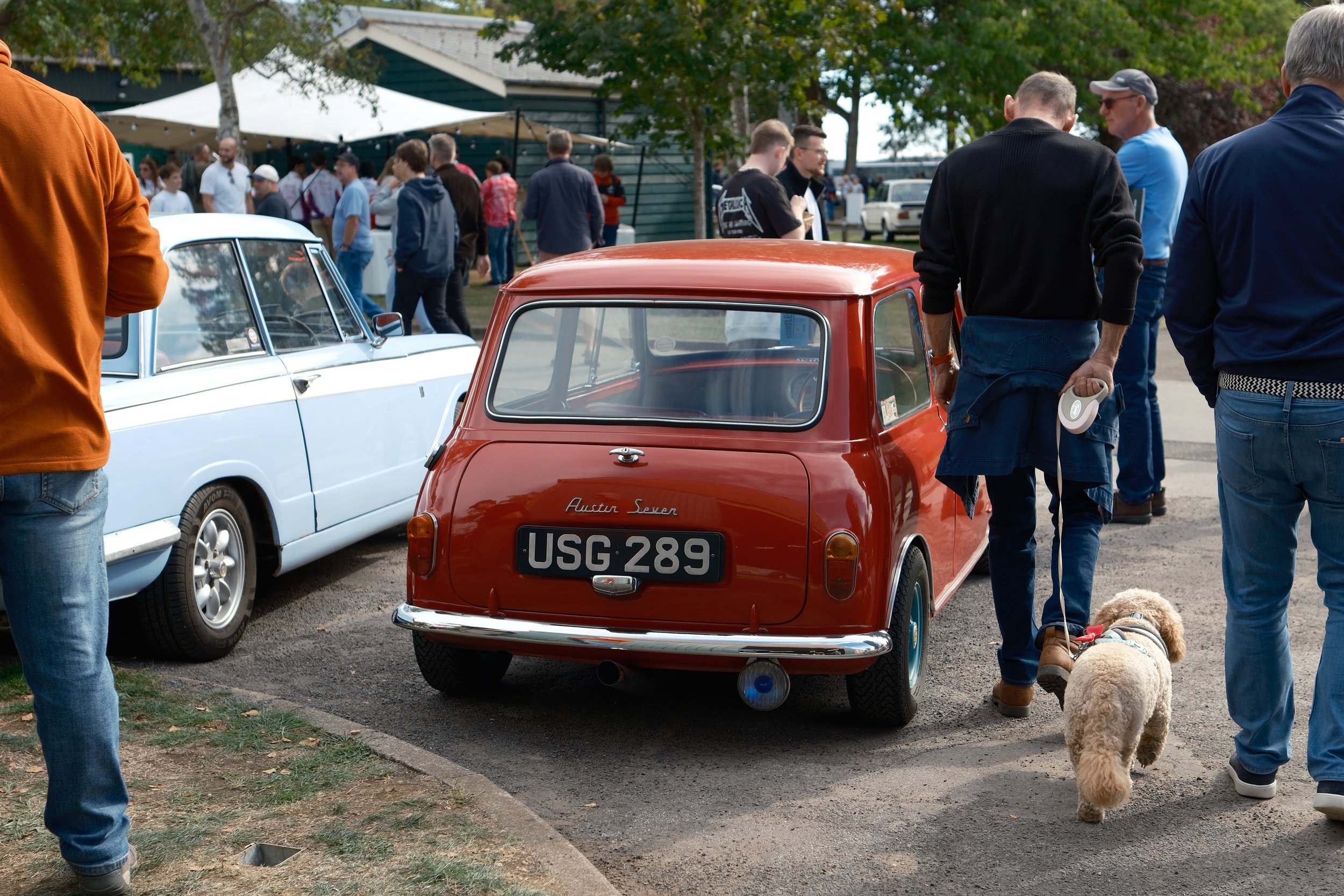 People attending a car show with vintage cars, including a red Austin Seven miniature car and a light blue classic car, in an outdoor setting with trees and a building in the background.