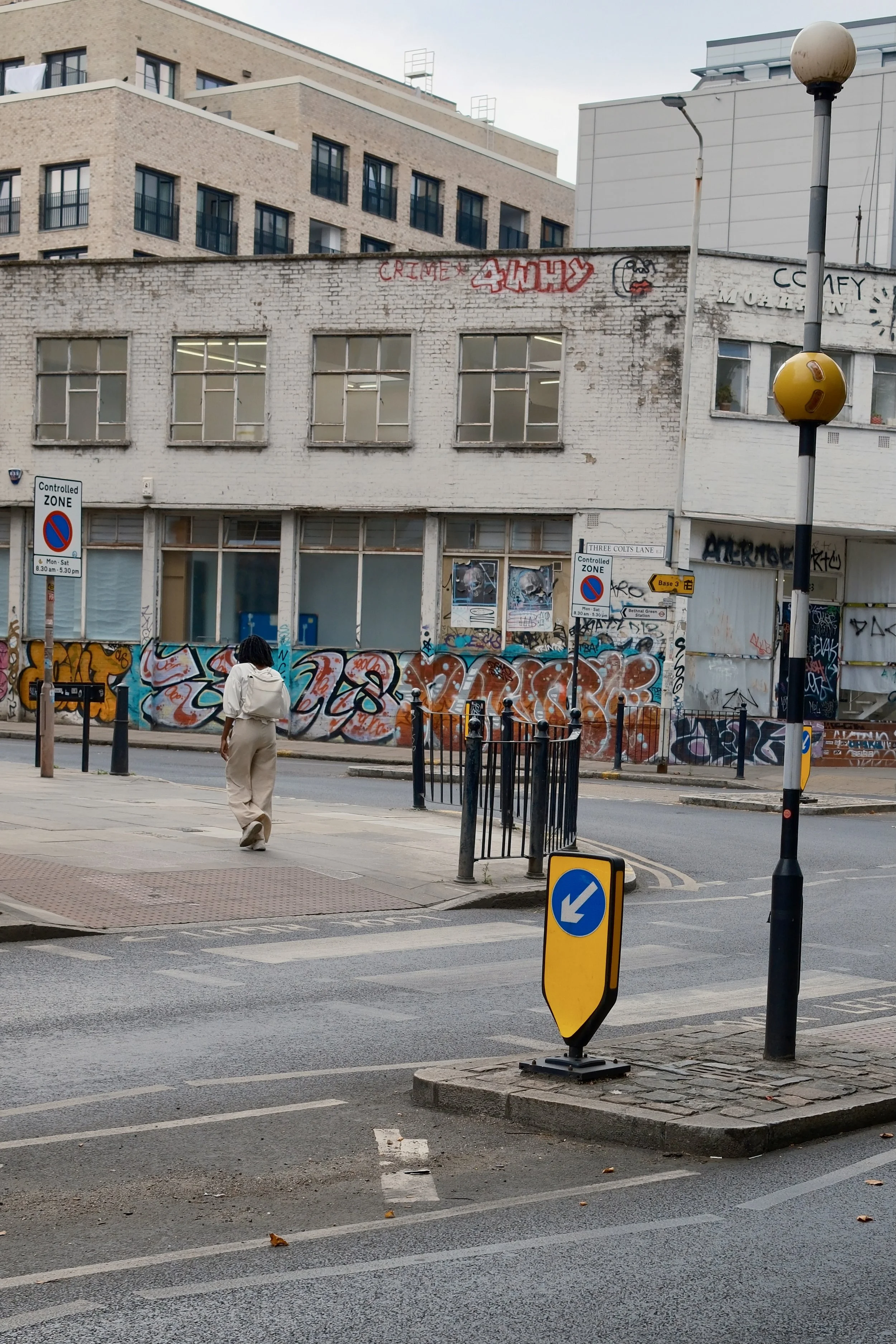 A person walking on a city sidewalk near a street with traffic signs, a black metal fence, and graffiti-covered walls. Buildings in the background.