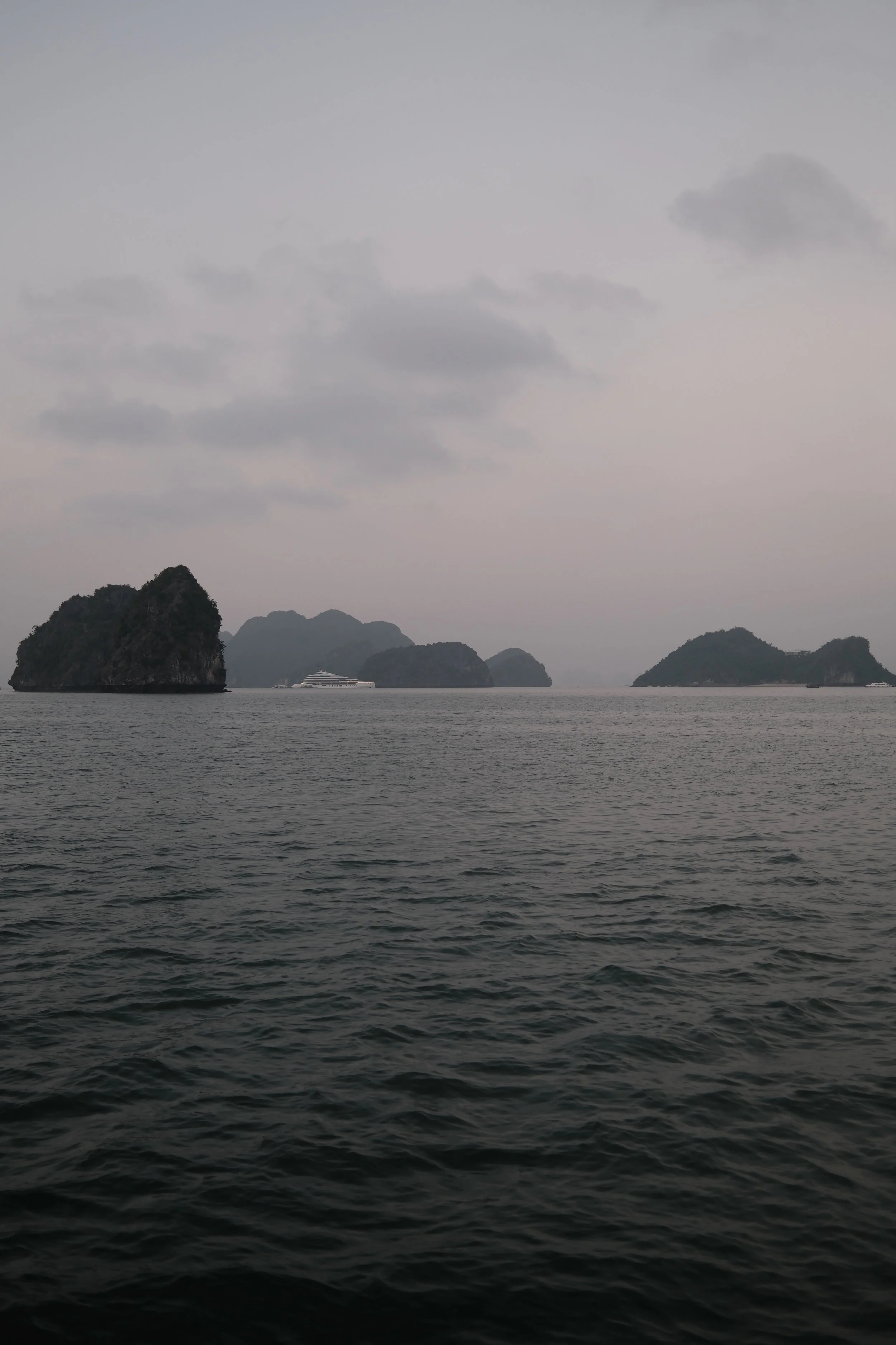 A seascape featuring a calm body of water in the foreground with several islands or large rocks visible in the distance. A white cruise ship is seen near the middle of the image between the islands. The sky above is overcast with grey clouds.