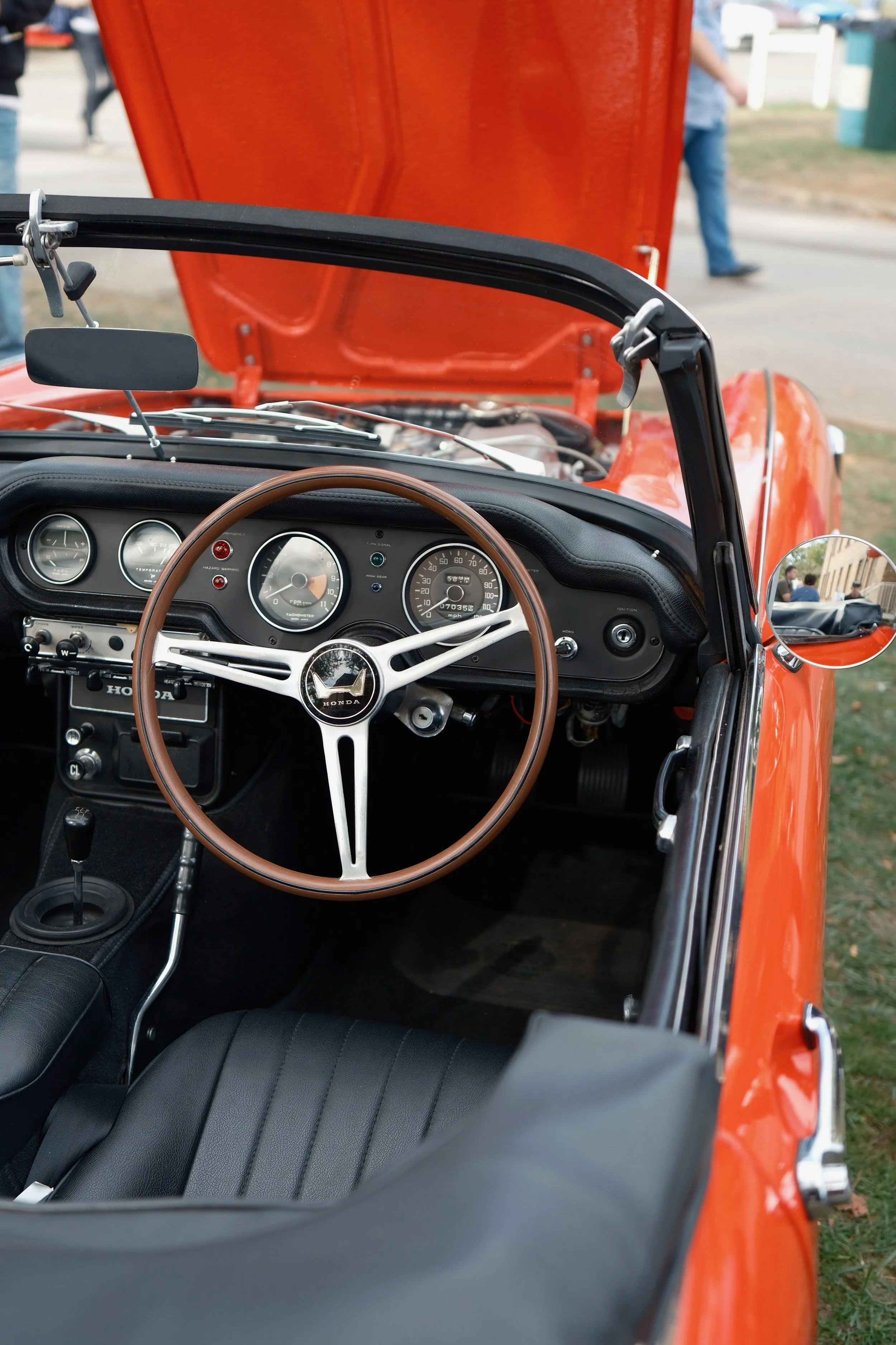 Interior of a vintage red convertible car with a wooden steering wheel, showing the dashboard with analog gauges and a gear shift, at an outdoor event with people in the background.