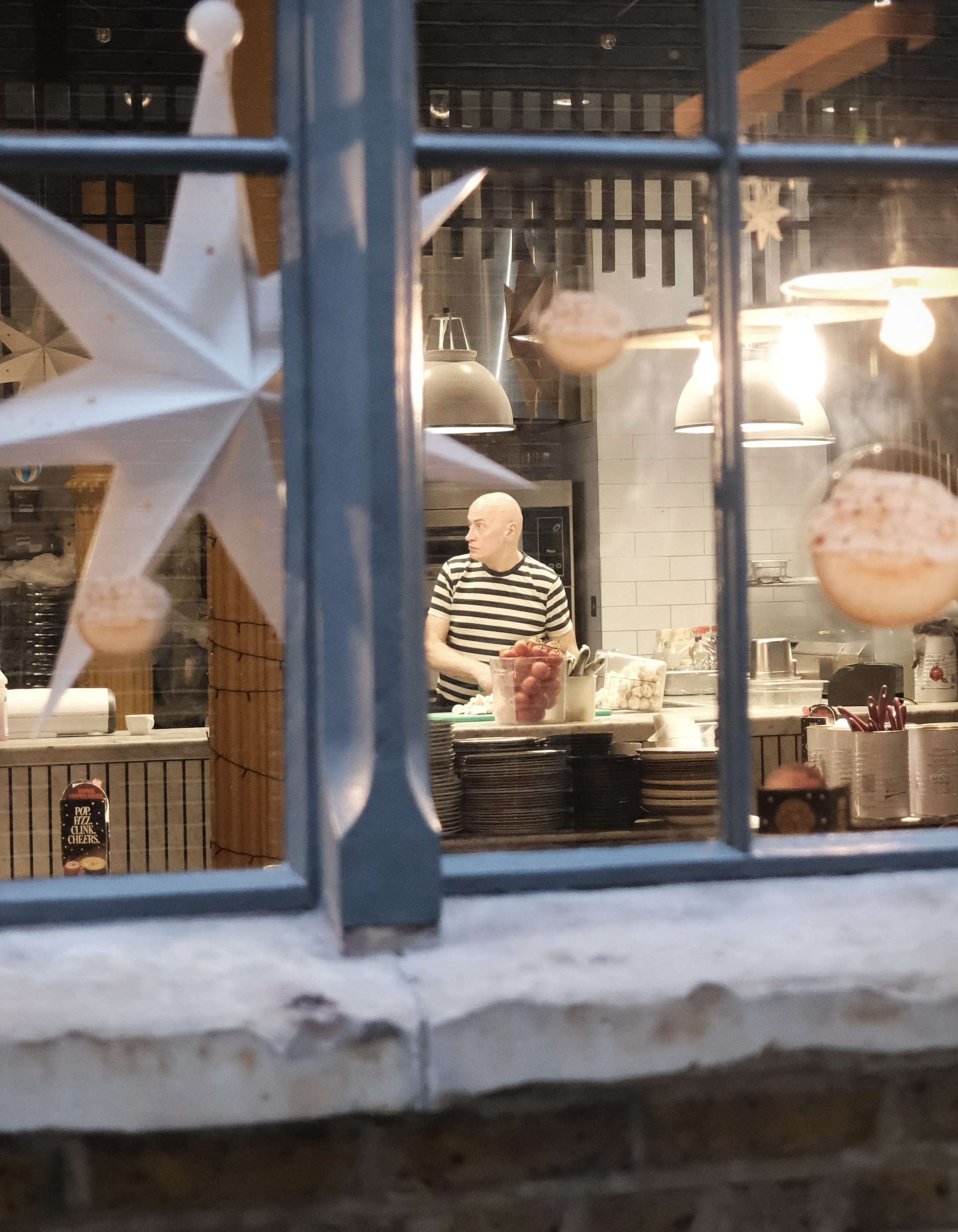 A man in a striped shirt preparing food in a kitchen, viewed through a window decorated with festive star and ball ornaments, with warm lighting overhead.