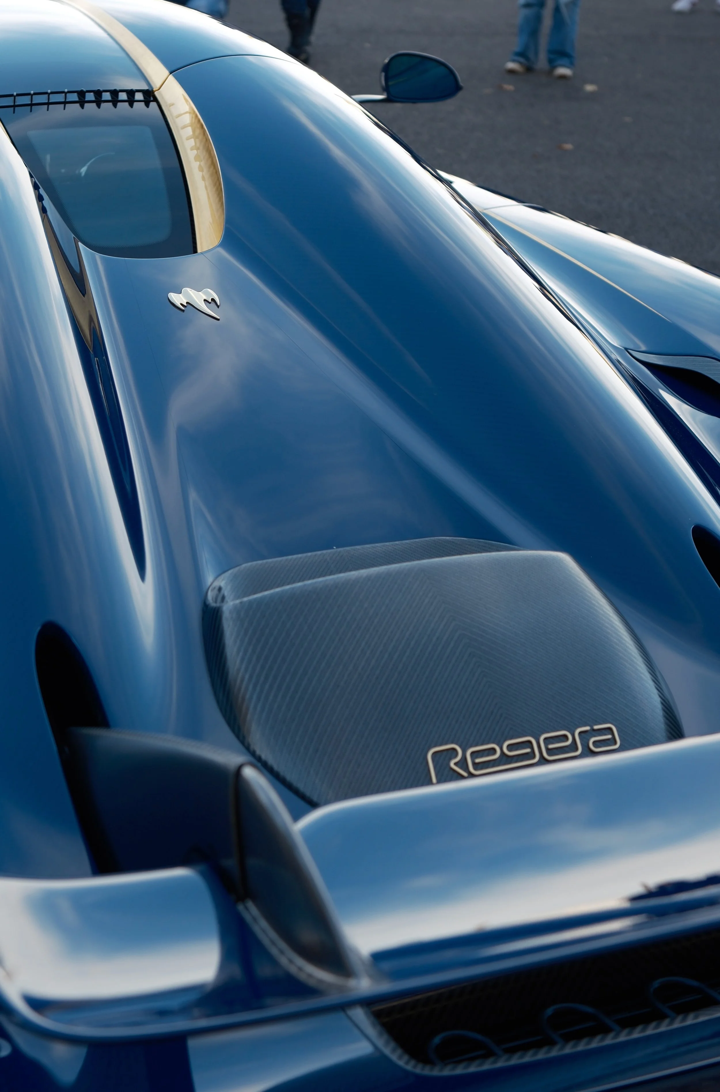 Close-up of a blue sports car with sleek design, showing the windshield, side mirror, and part of the hood with the 'Regera' badge. The car has a reflective surface and carbon fiber accents.