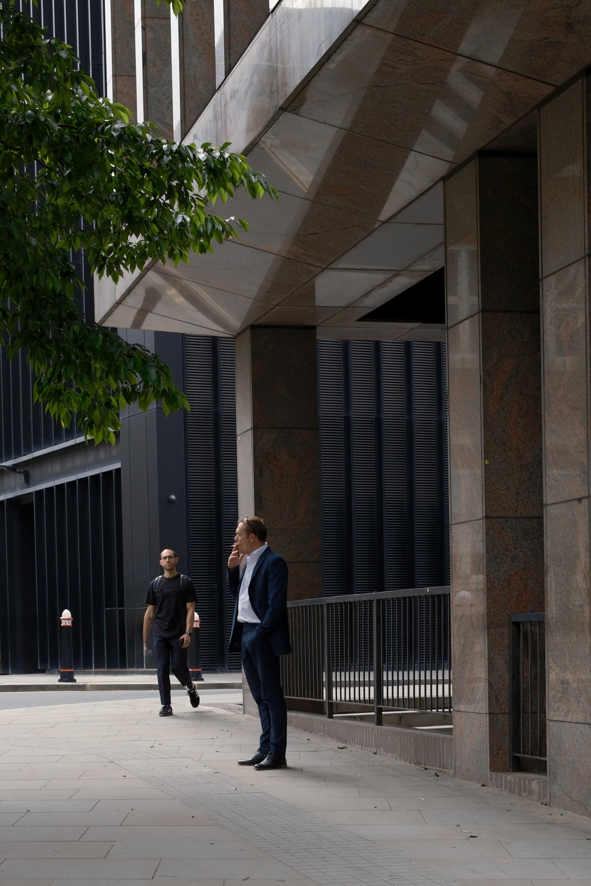 Two men walking outside a modern building with reflective glass and stone walls, one in a business suit talking on a cell phone and the other in casual attire carrying a backpack.