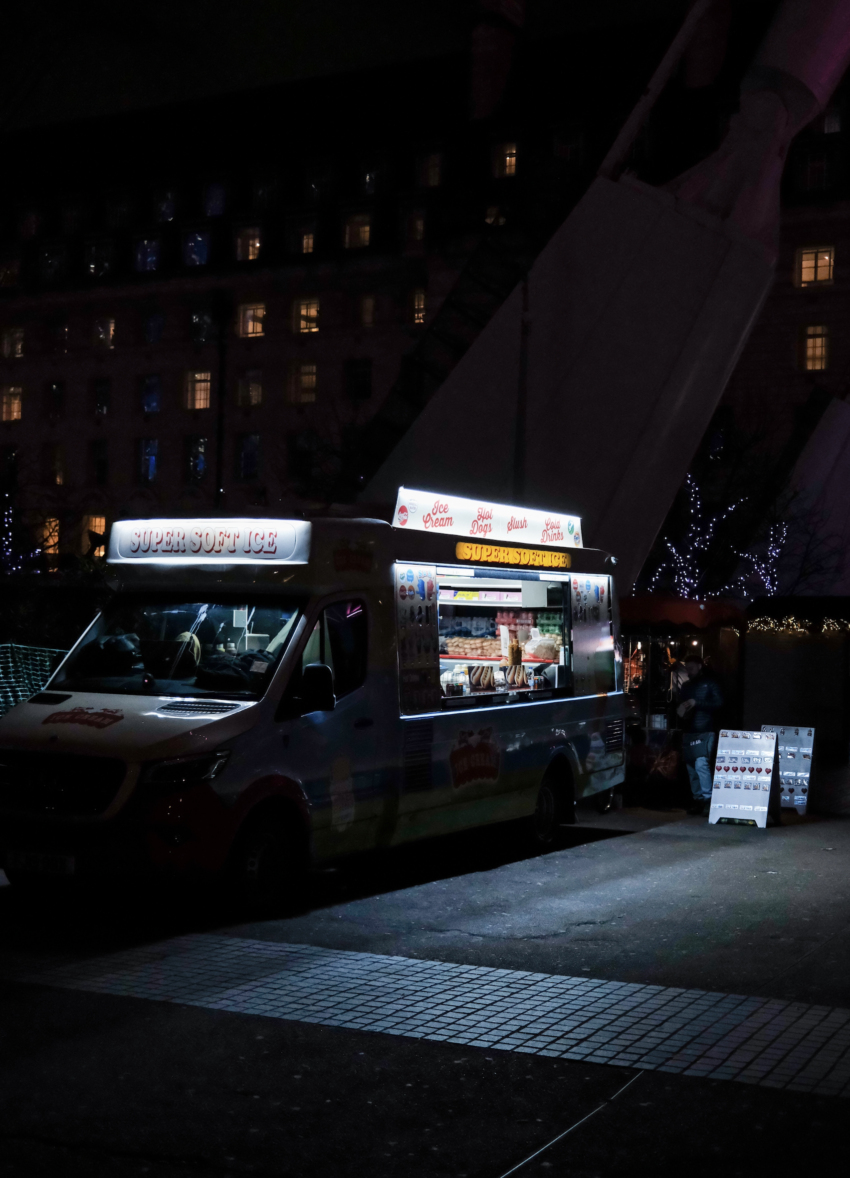 Night scene of a food truck named 'Super Soft Ice' stationed on a city street, with a building and window lights in the background and a menu board on the sidewalk.
