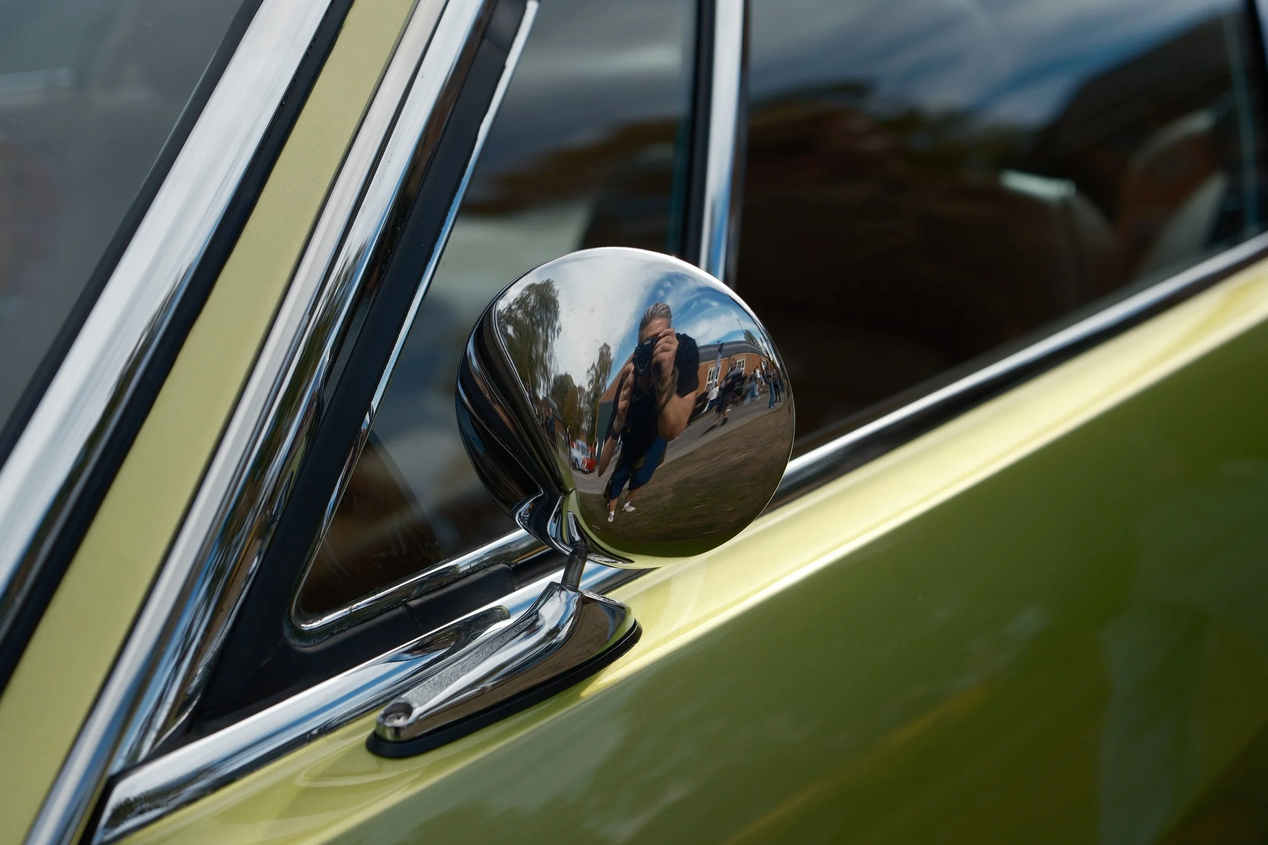 Close-up of a reflective side mirror on a vintage car, showing the person taking the photo, trees, and a building in the reflection.