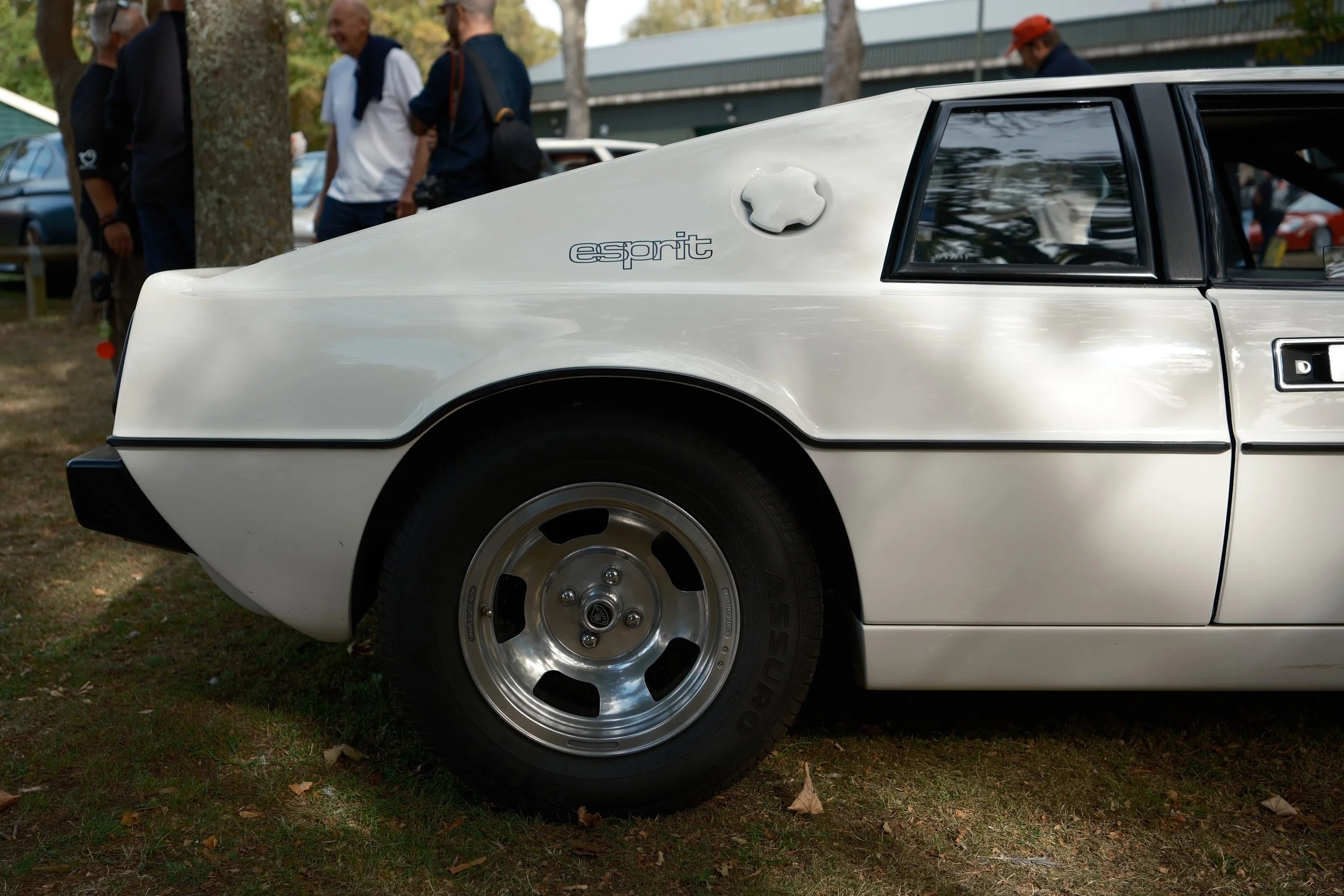 Close-up of a white classic car's front side with a black rubber tire, 'esprit' badge, and a sleek black window. In the background, people are gathered outdoors near trees and other vintage cars, indicating a car show or meetup.