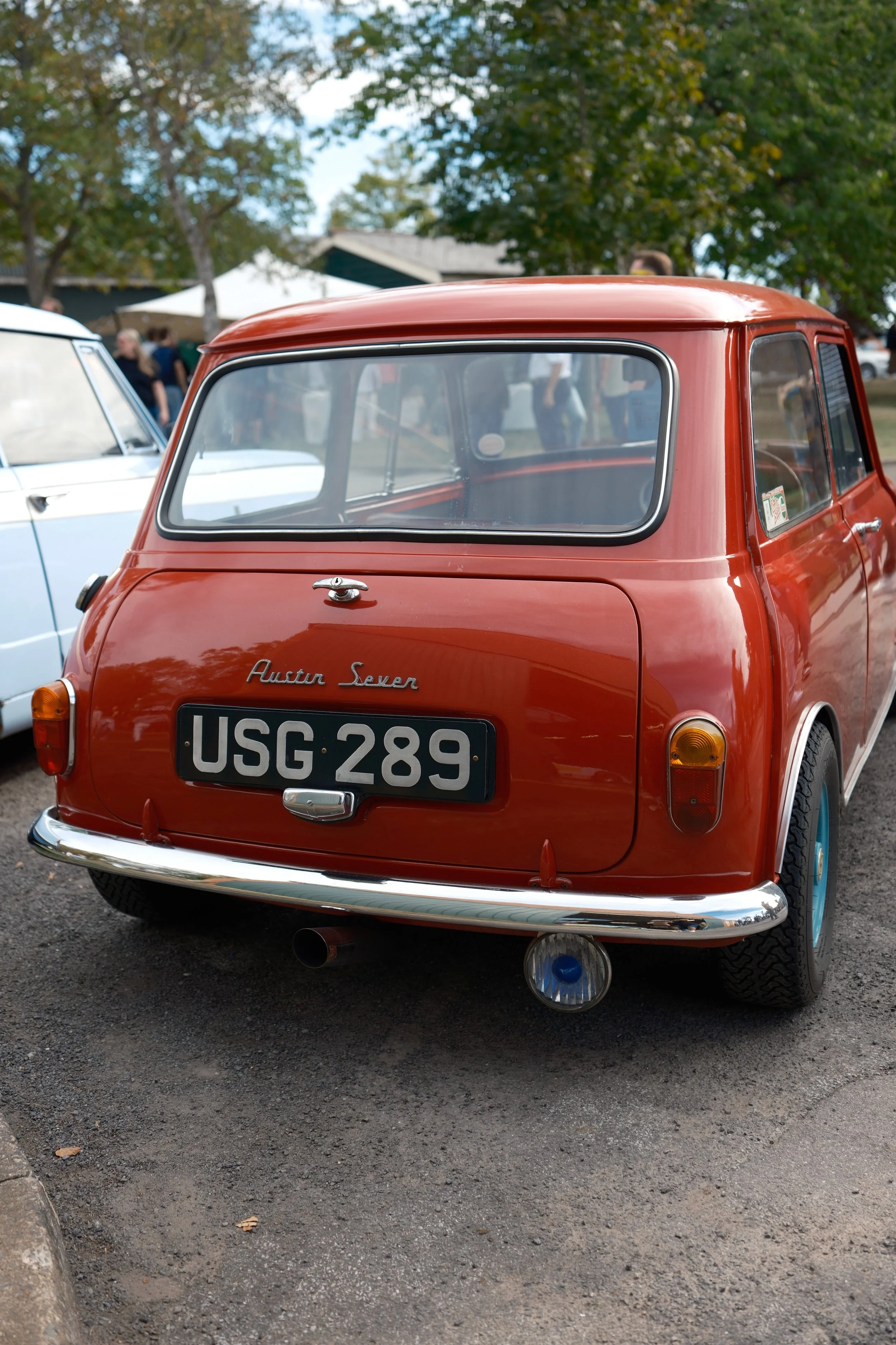 Rear view of a vintage red Austin Seven microcar with a custom license plate 'USG 289' parked on a gravel surface, with trees and people in the background.