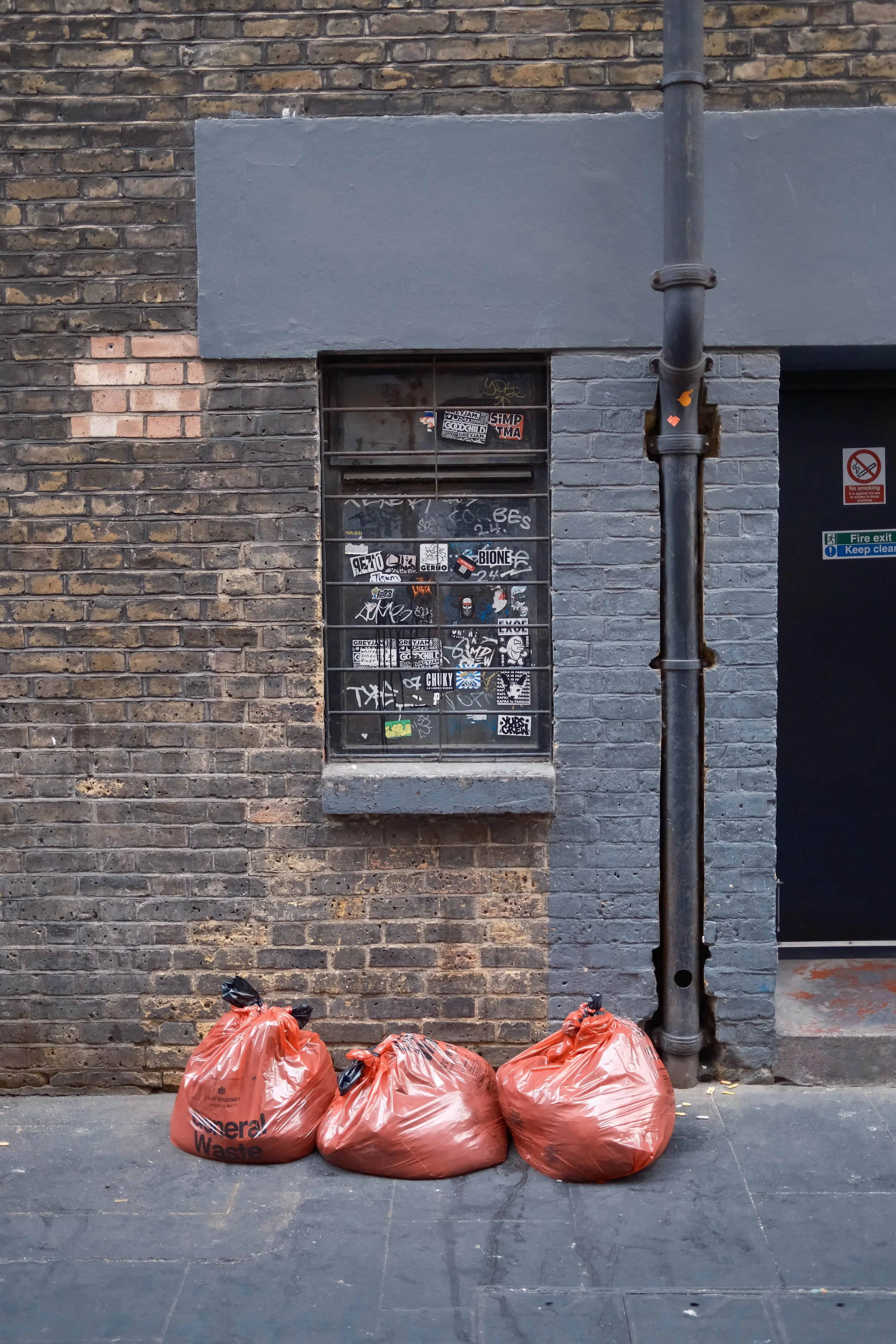 Five orange trash bags labeled 'General Waste' placed on a sidewalk in front of a brick wall with a window and grey painted section, next to a drainpipe.