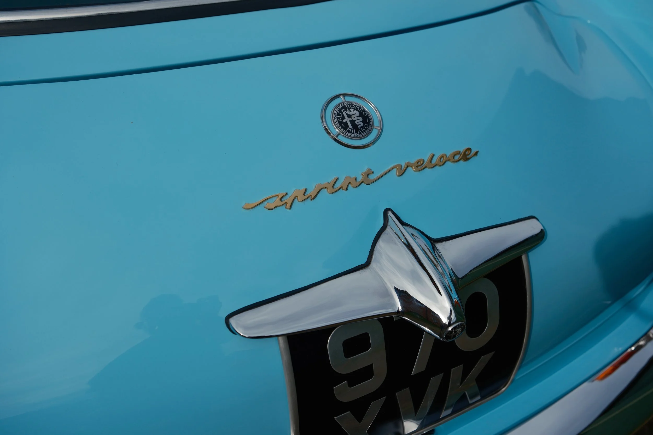 Close-up of the front of a light blue Alfa Romeo car, featuring the emblem and the 'Spider Veloce' badge and part of the grille and hood.