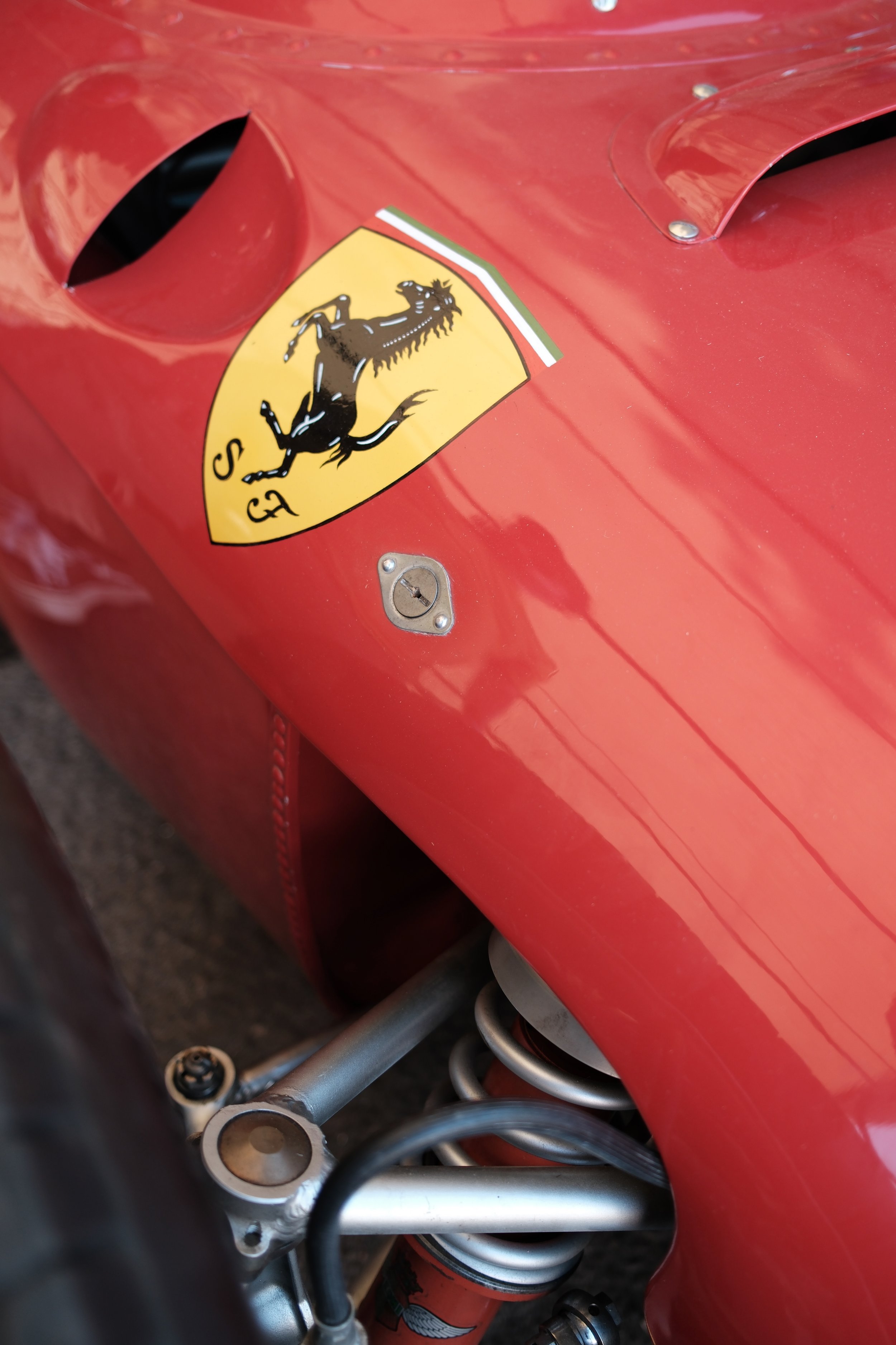 Close-up of a red motorcycle with a Ferrari logo sticker on the fuel tank, showing mechanical parts such as metal tubing and a shock absorber.