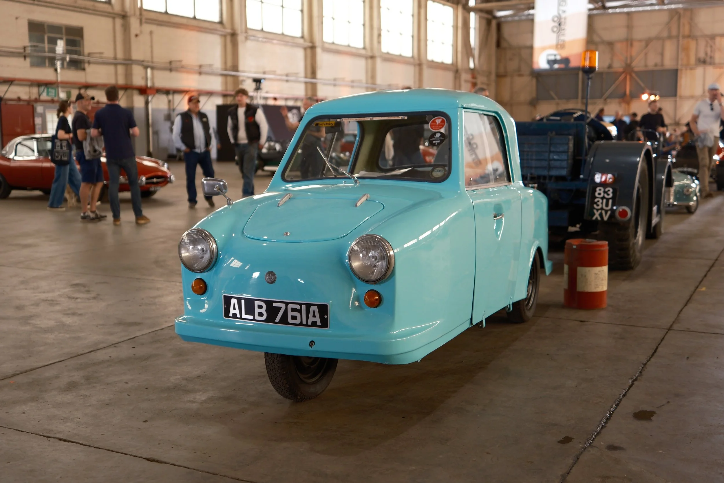 A small, vintage, blue three-wheeled vehicle with the license plate ALB 76IA, displayed at an indoor auto show, with several people and classic cars in the background.