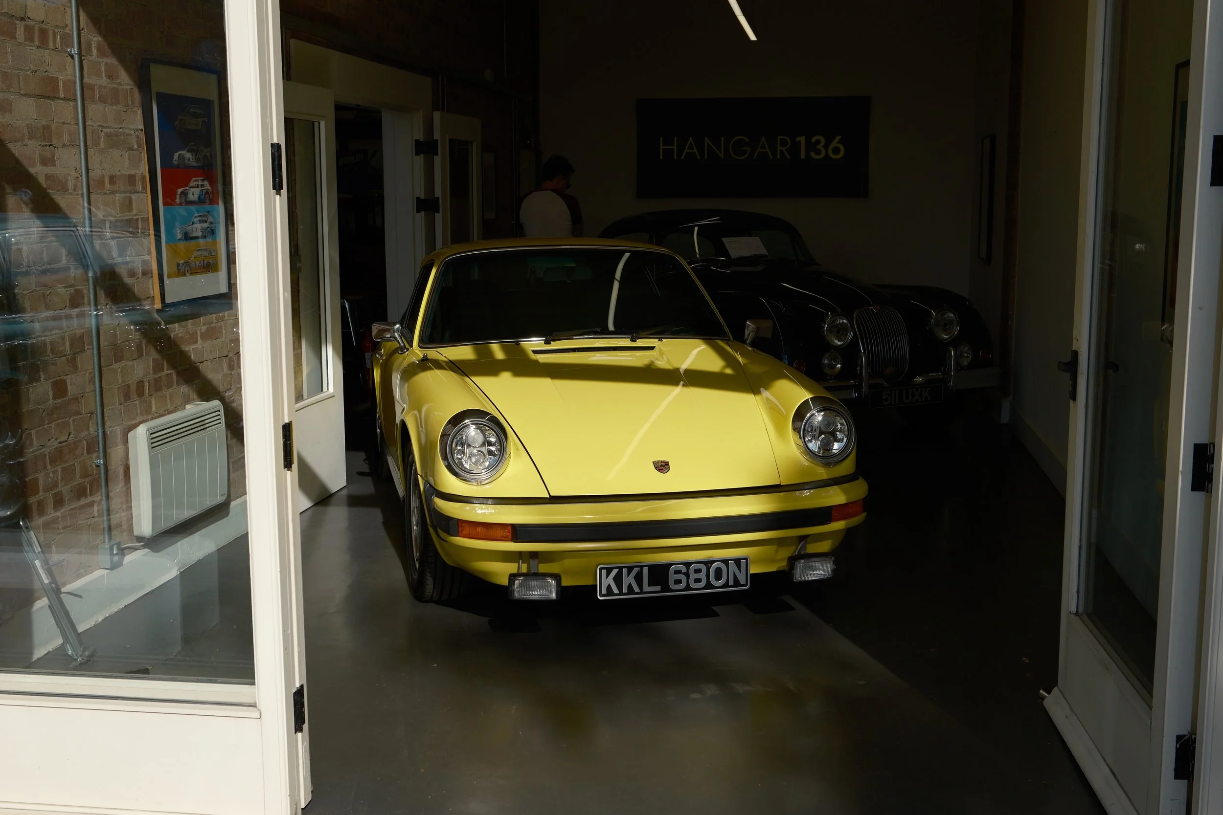 Yellow classic Porsche car parked indoors, with a black classic car behind it, inside a showroom or garage labeled 'HANGAR 136'.