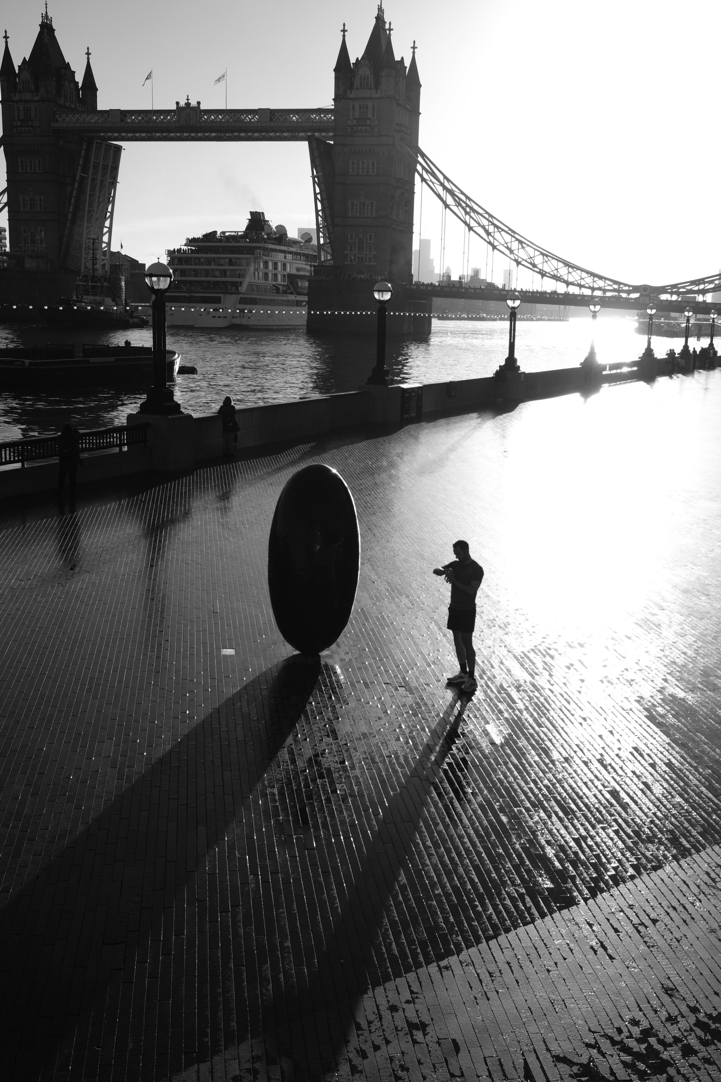 Black and white photo of Tower Bridge in London, with a large cruise ship docked underneath, and a person standing by a sculpture on the waterfront walkway, casting long shadow.