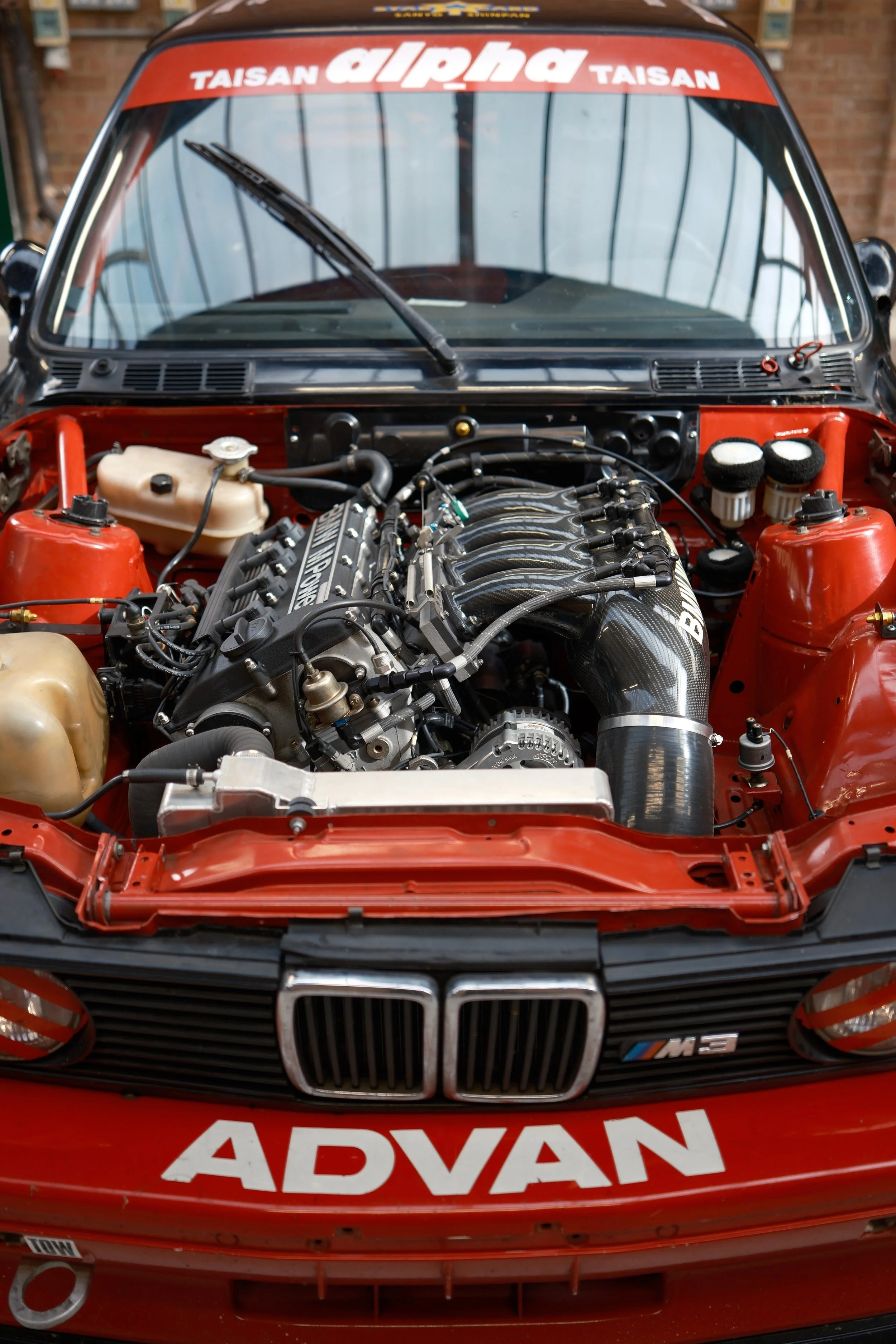 Front view of a red BMW race car with the engine exposed, showing a high-performance engine and various car stickers including 'Alpina', 'Taisan', and 'Advance'.