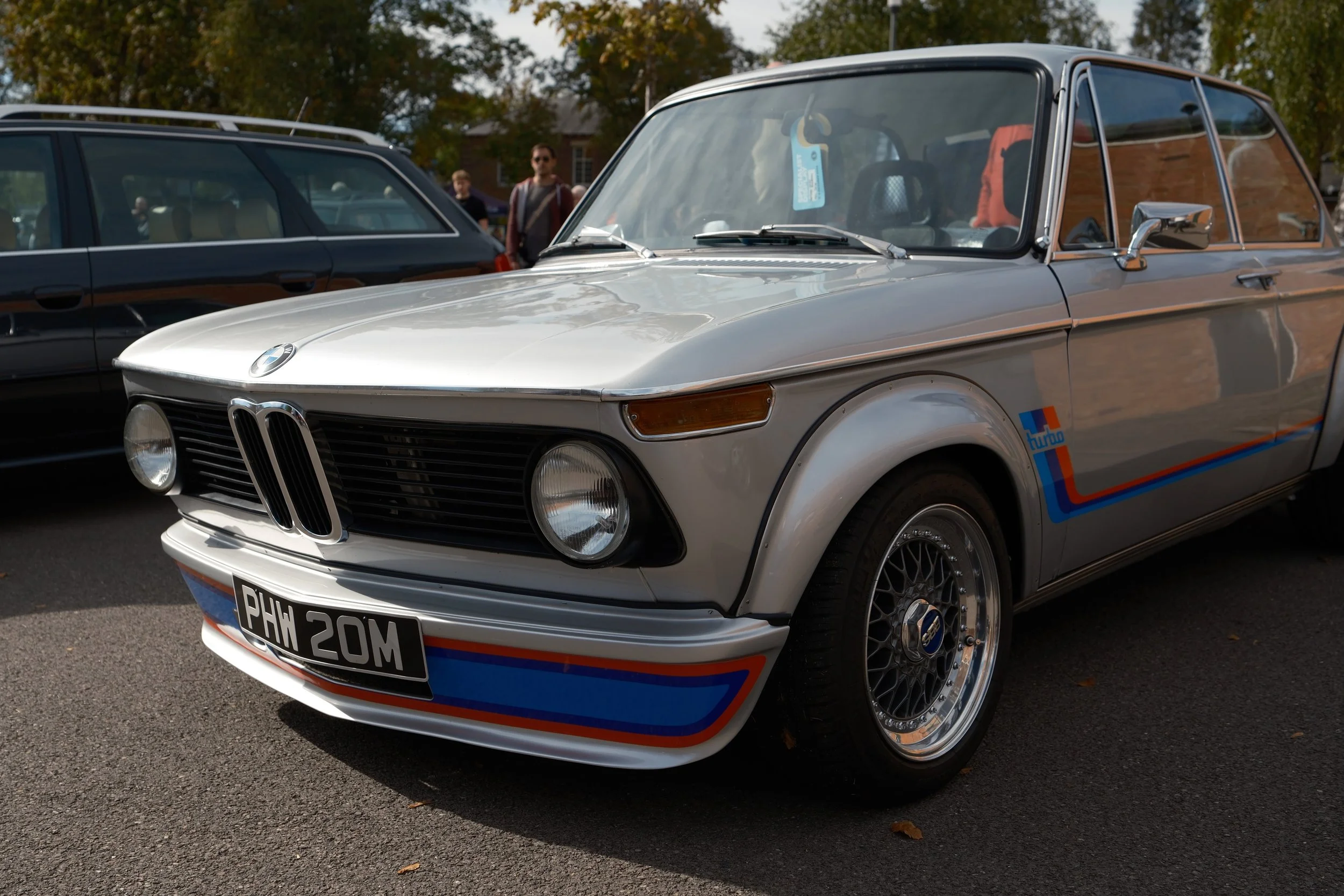 A vintage silver BMW car with racing decals parked outdoors, with people and trees in the background.