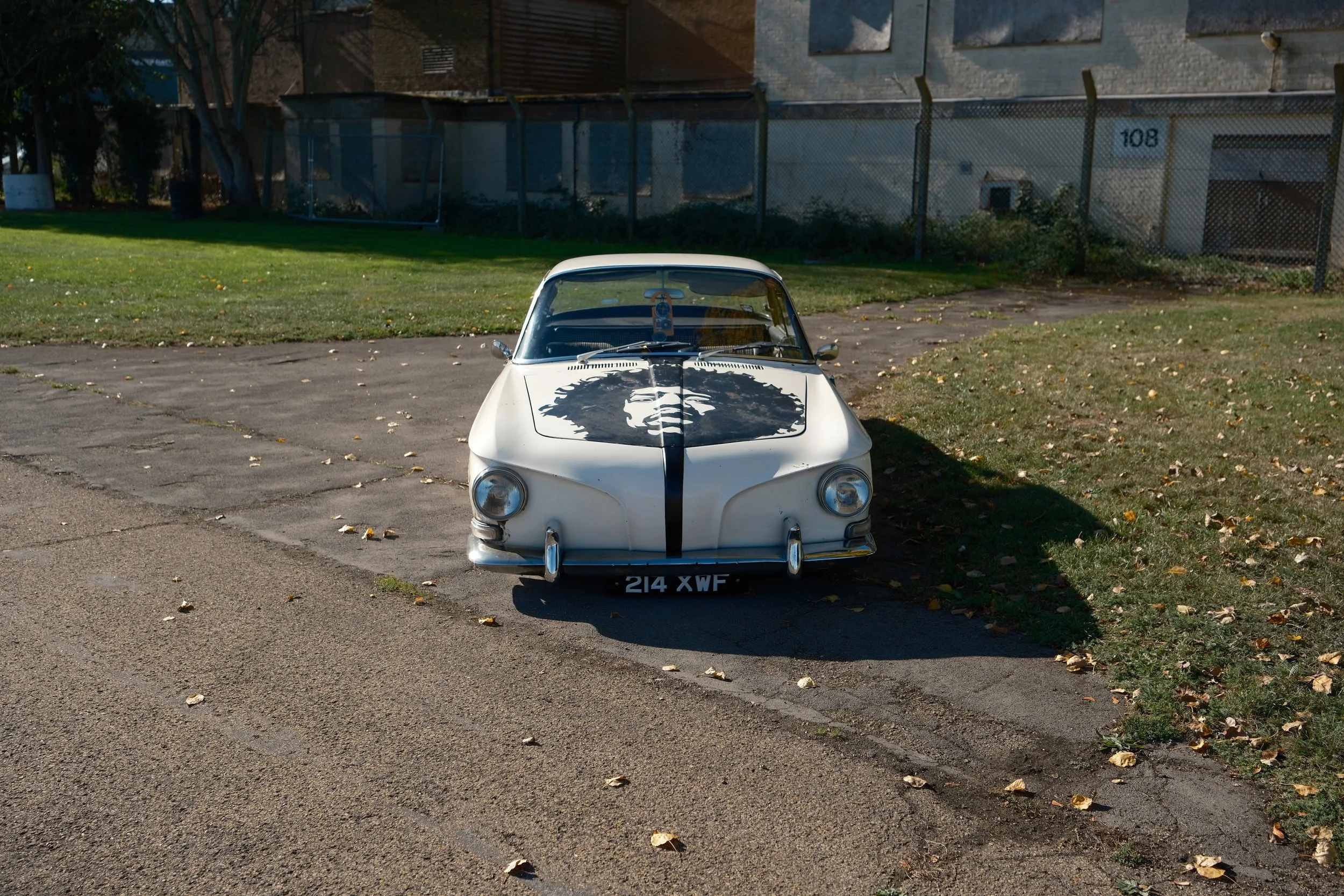 A vintage white car with a lion face graphic on the hood is parked in a lot with a grassy area and a chain-link fence in the background.