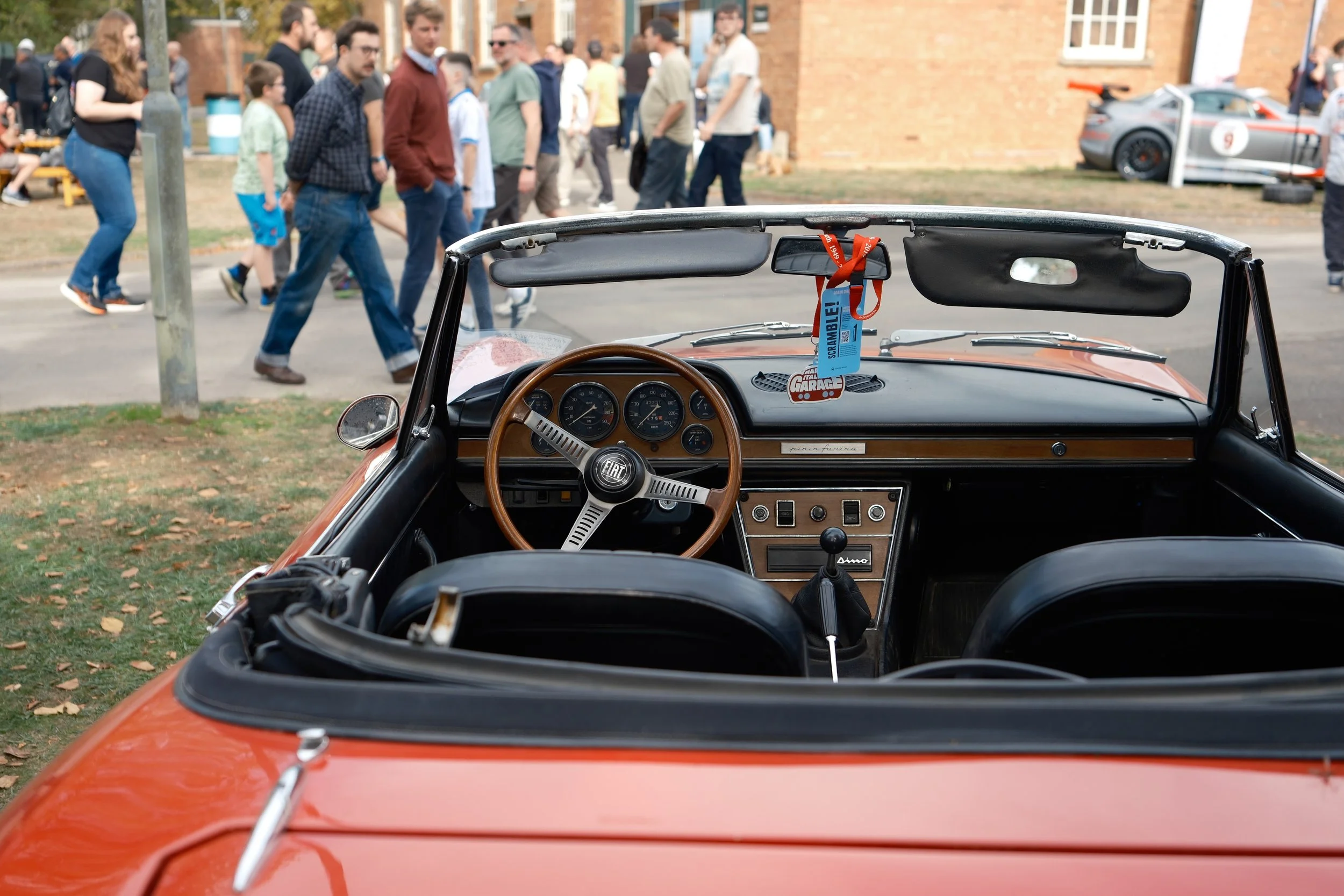 The interior of a vintage Fiat car with a wooden steering wheel, dashboard gauges, and a gear shifter, viewed from the windshield, with a crowd of people walking by outside in an outdoor event.
