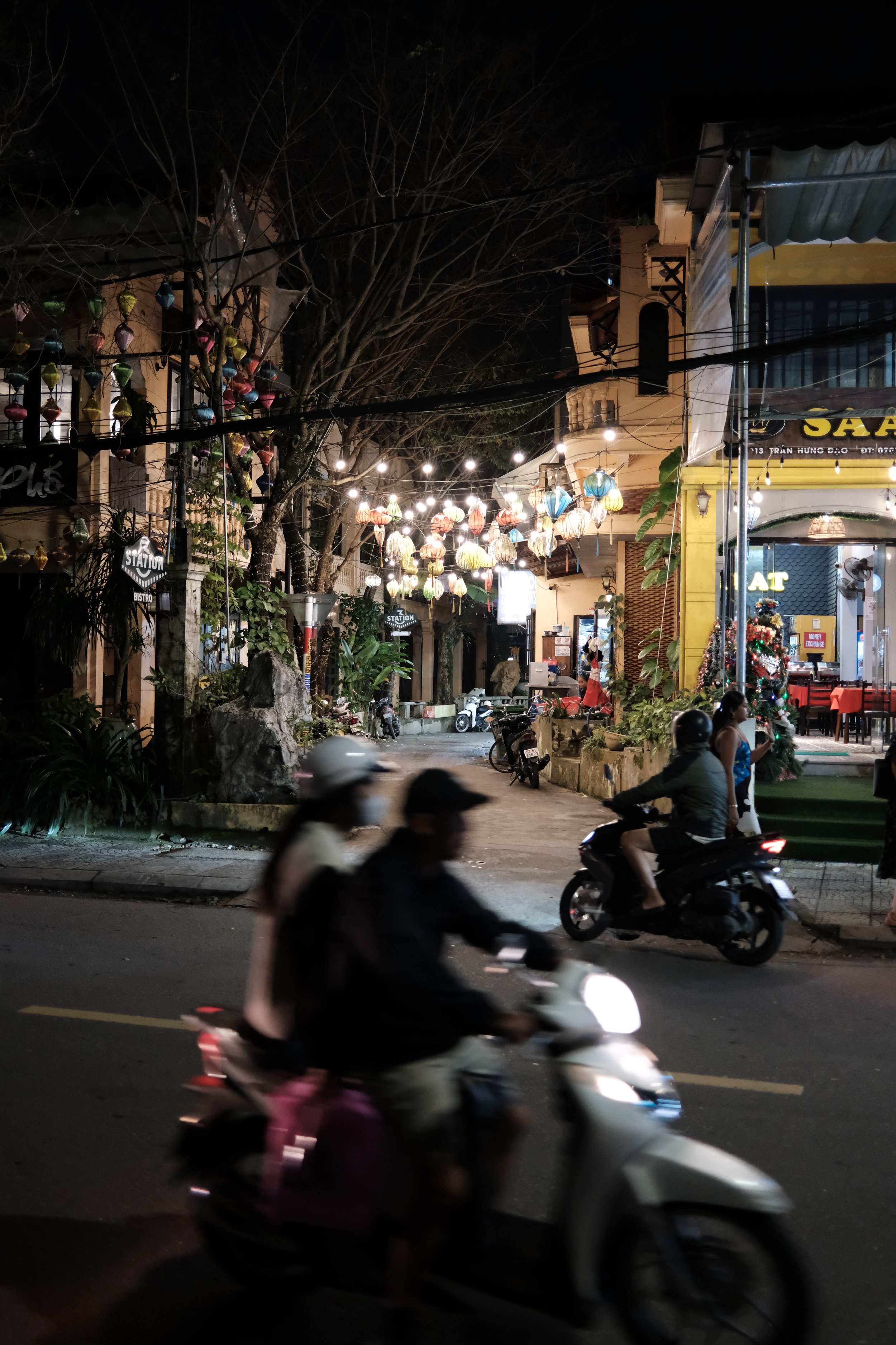 Night street scene with colorful hanging lanterns, people riding scooters, and illuminated storefronts.