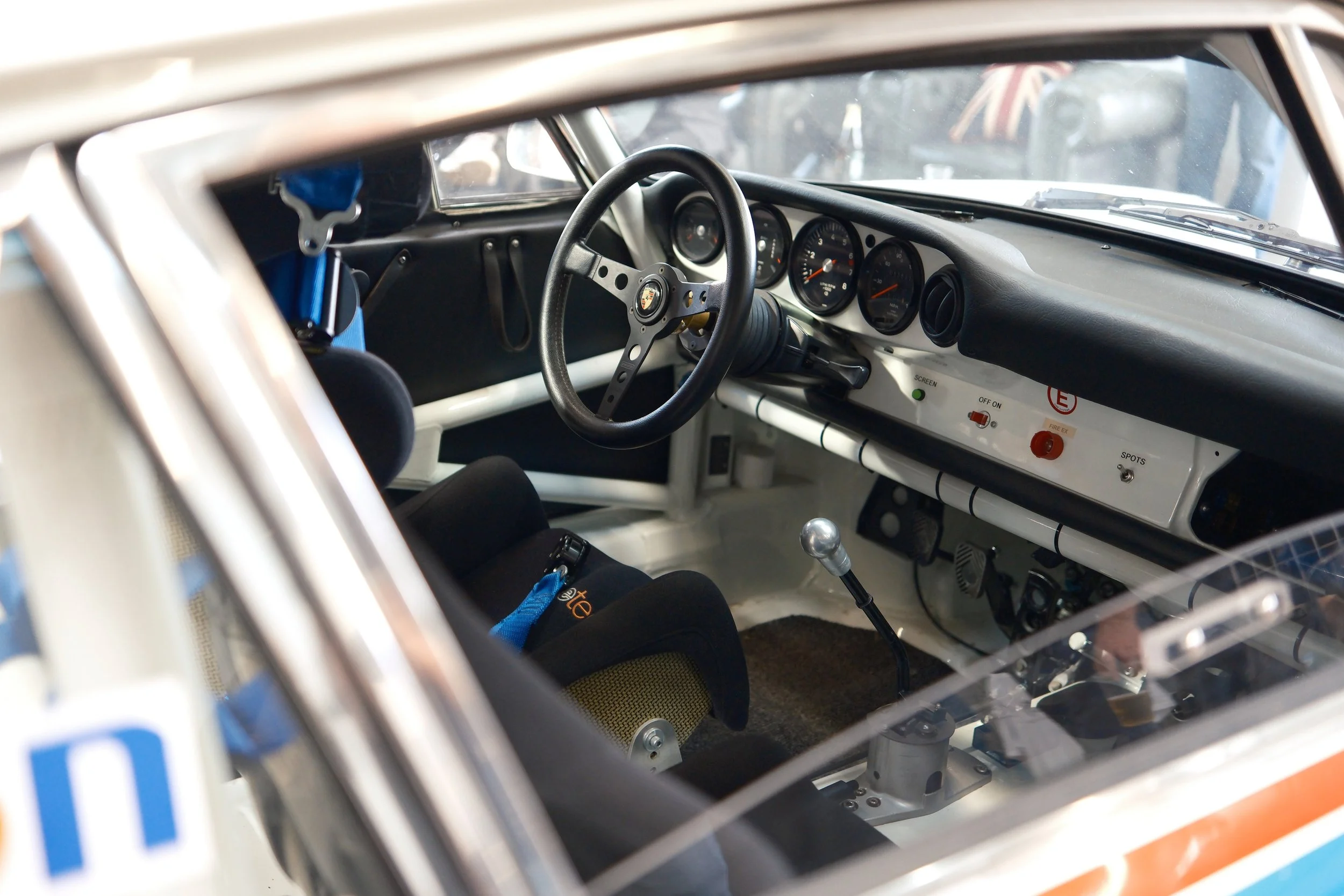 Interior of a race car showing the steering wheel, dashboard gauges, gear shift, and seat with harnesses.