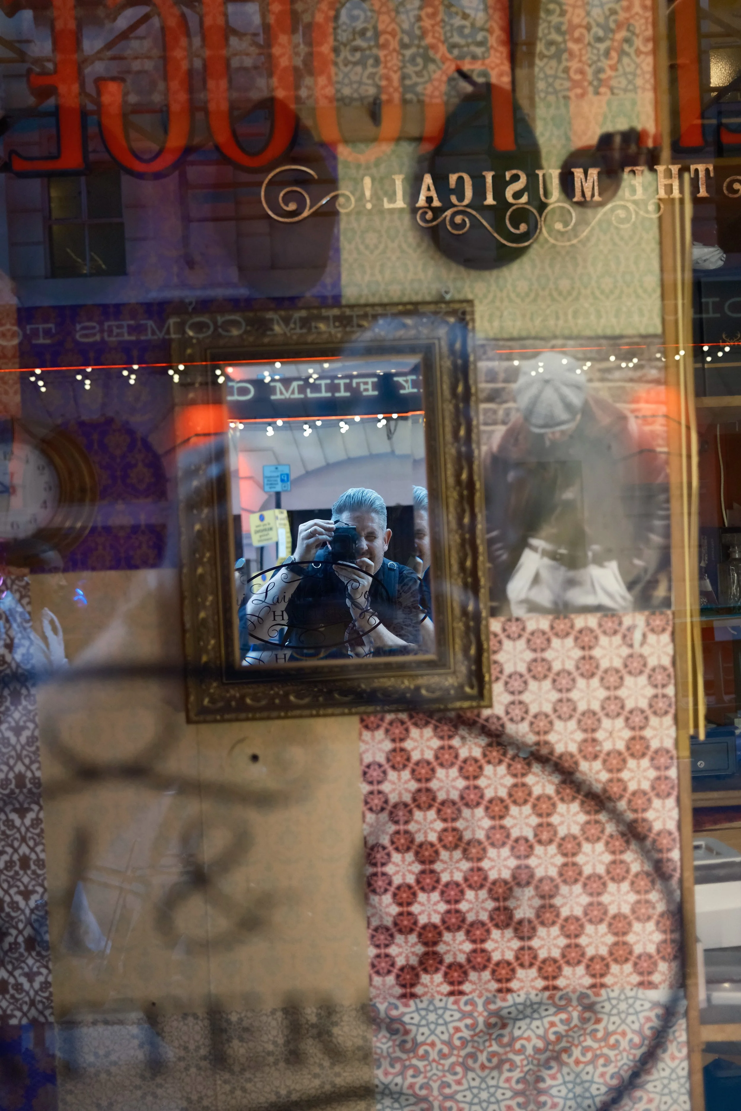 Reflection of a person taking a photo with a camera, seen in a glass mirror which has decorative text and ornate picture frames. The setting is a vintage or antique shop with patterned wallpaper and tile flooring.