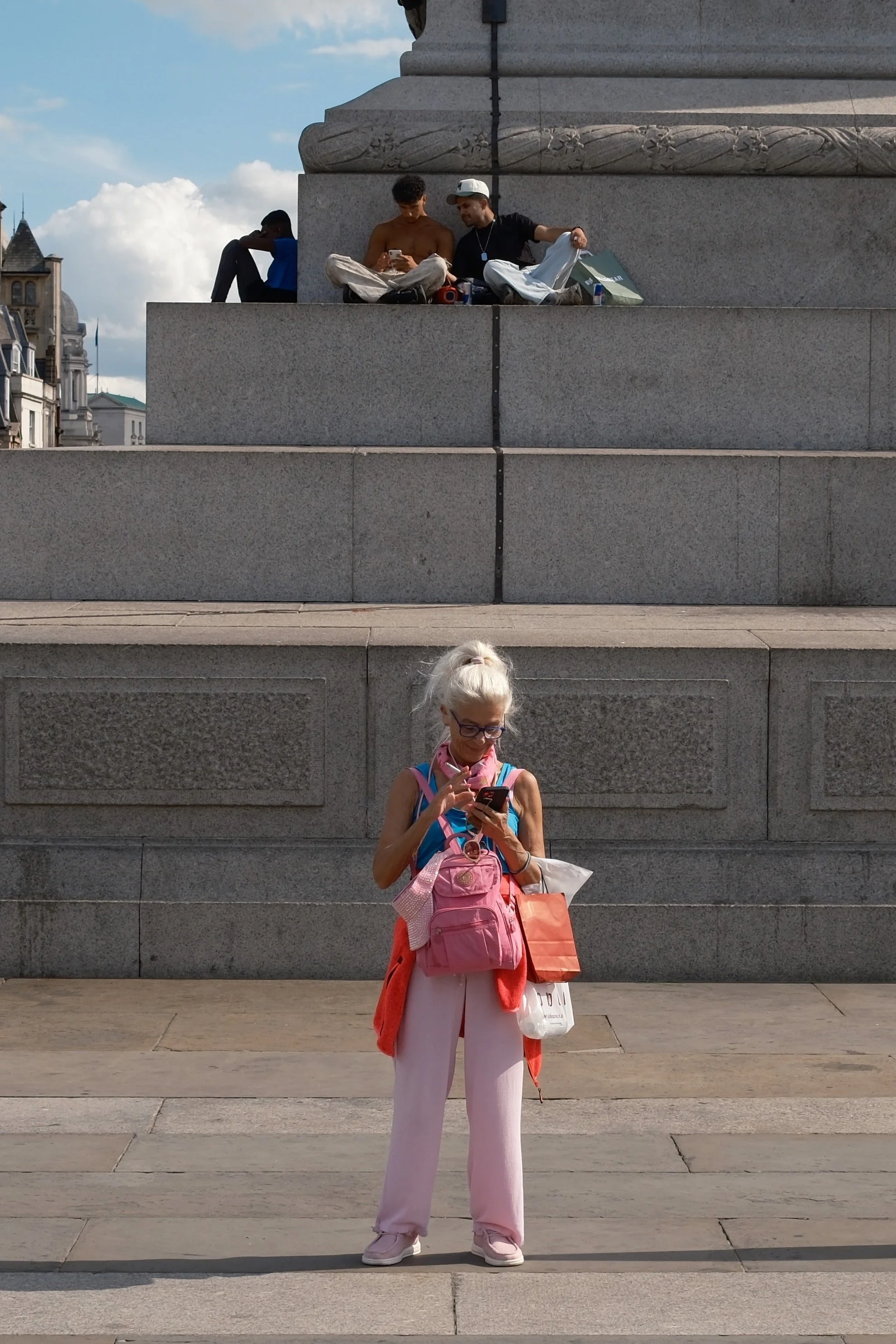 A woman with white hair, wearing glasses, a pink backpack, and pink and white clothing, stands in front of a stone wall looking at her phone. In the background, two young men sit on a ledge, one shirtless and the other wearing a black shirt and white