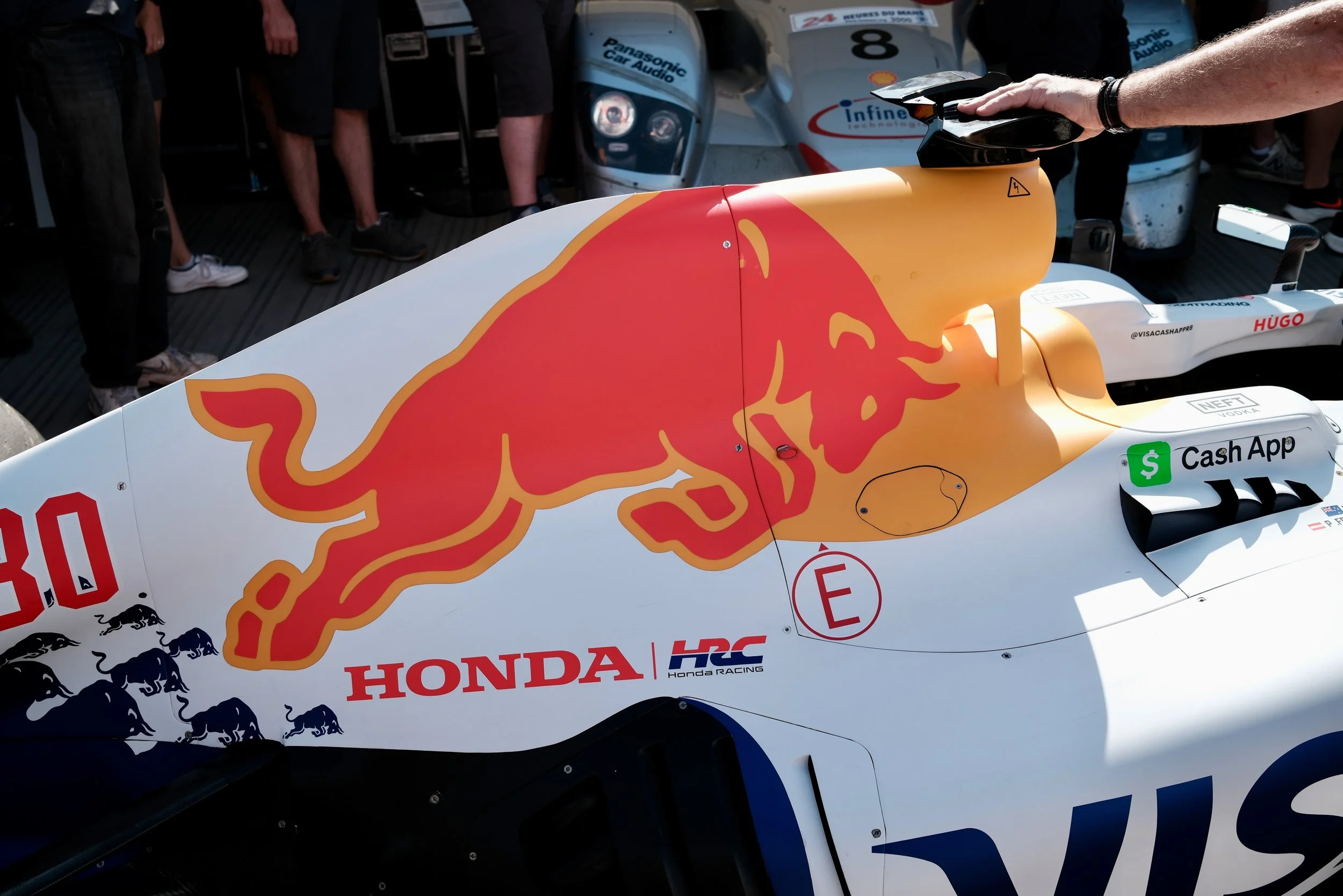 Close-up of a Formula 1 race car with a red bull logo and other sponsor logos, surrounded by people, with parts of other race cars visible in the background.