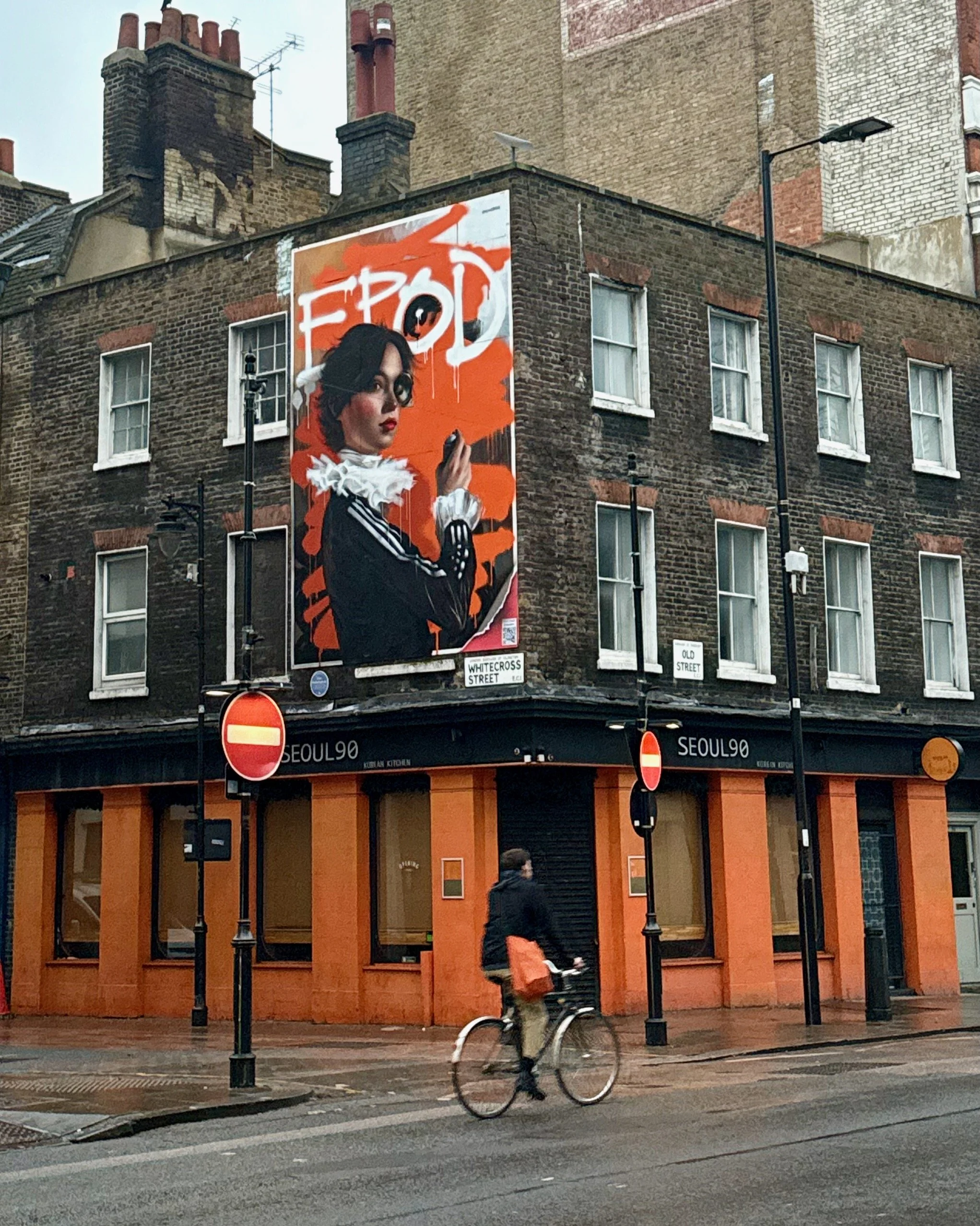 A large building with a brick facade. There is a prominent poster on the building showing a woman with dark hair holding a cigarette, with the word 'F*CK'. A cyclist rides past in the foreground, and there are street signs reading 'Whitecross Street'