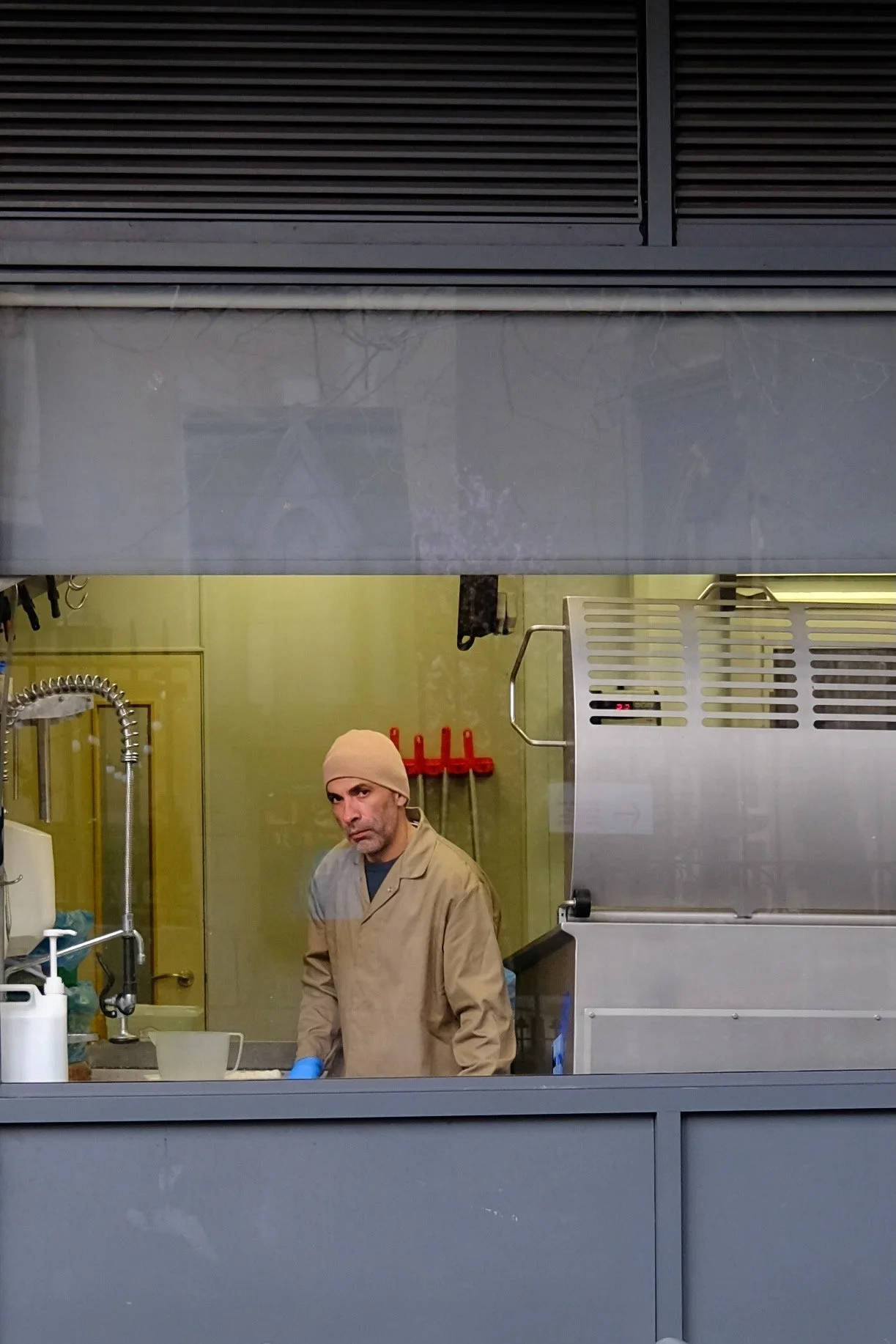 A man in a beige coat and beanie stands in a kitchen behind a glass window, with a yellow wall and kitchen equipment visible. He is looking at the camera with a serious expression.