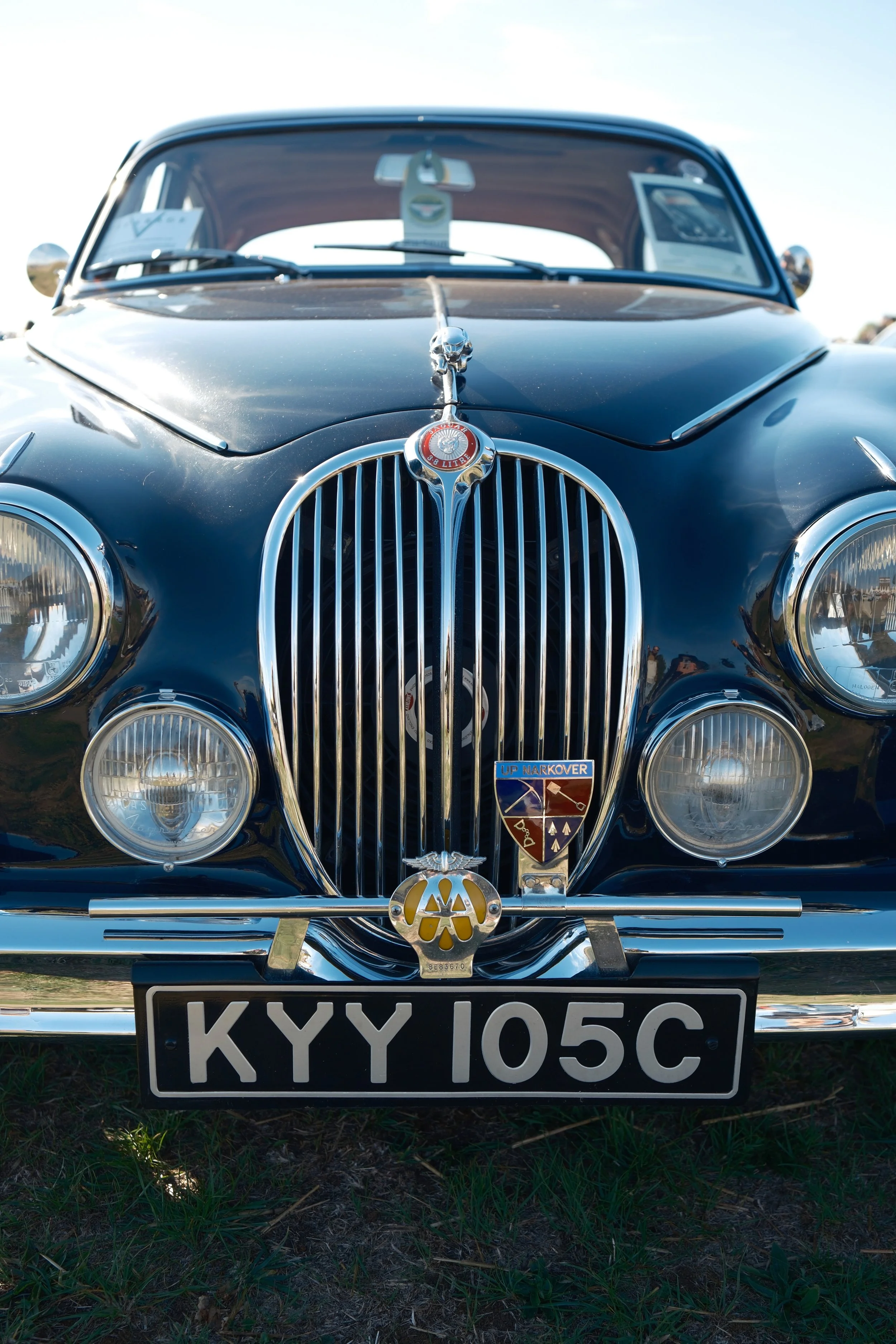 Front view of a vintage black car with mail markers, badges, and a black license plate with white letters reading 'KXY 105C,' parked on grass under a clear sky.