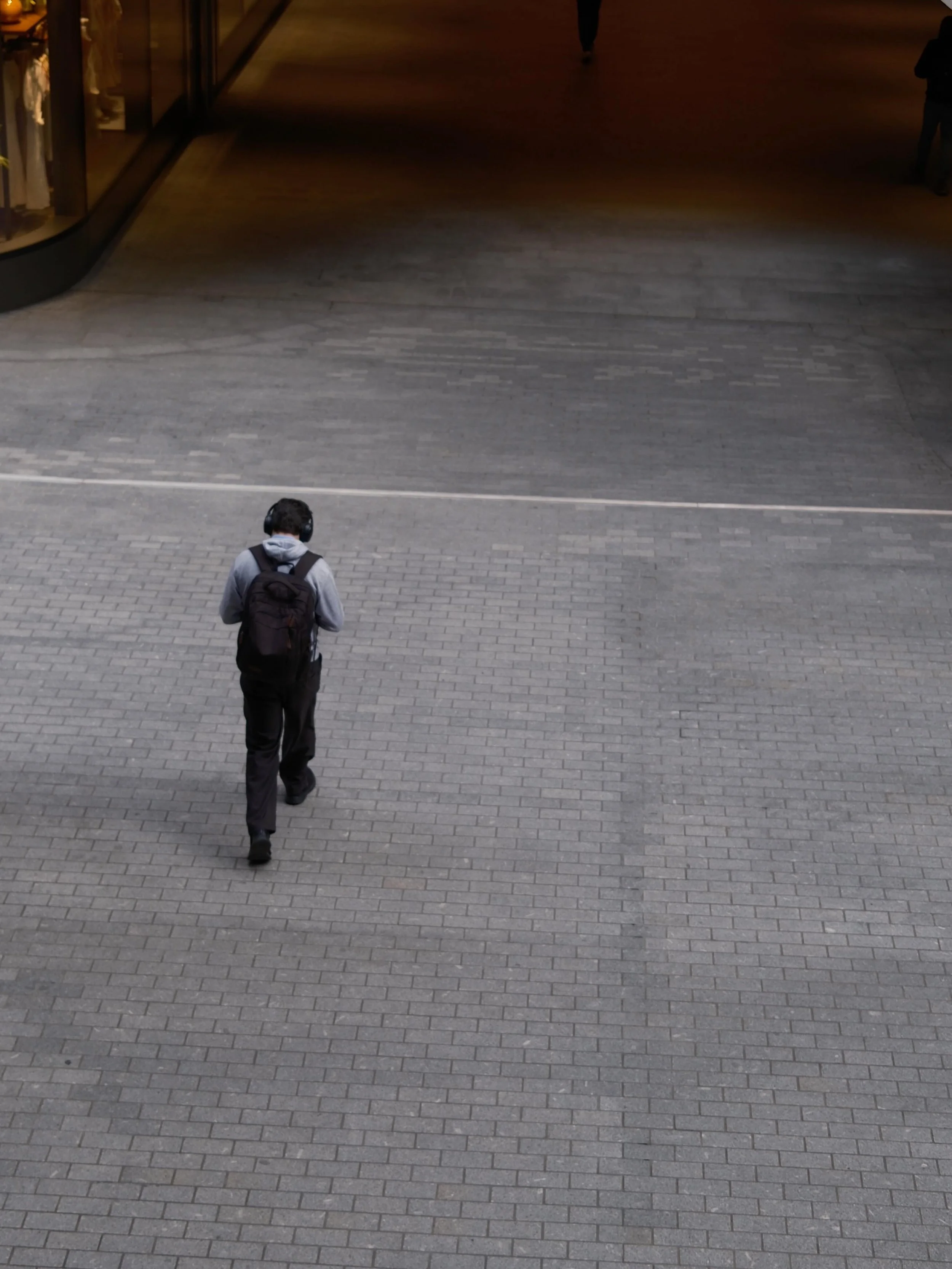 A person walking on a paved walkway near a shopping mall entrance, wearing a backpack and headphones.