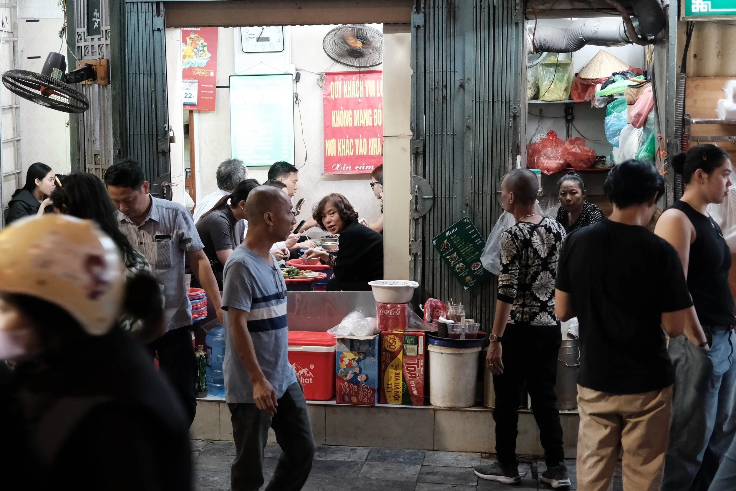 Crowded street food stall in Vietnam with people waiting and a sign in Vietnamese.