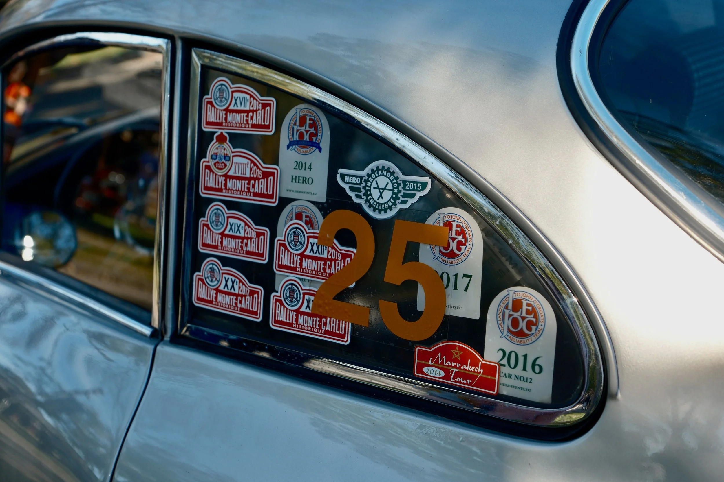 Close-up of a car window with various stickers from rally events and adventures, including Rallye Monte-Carlo and Marrakeh Tour, along with a large orange number 3 and a partial view of a car's blue interior.