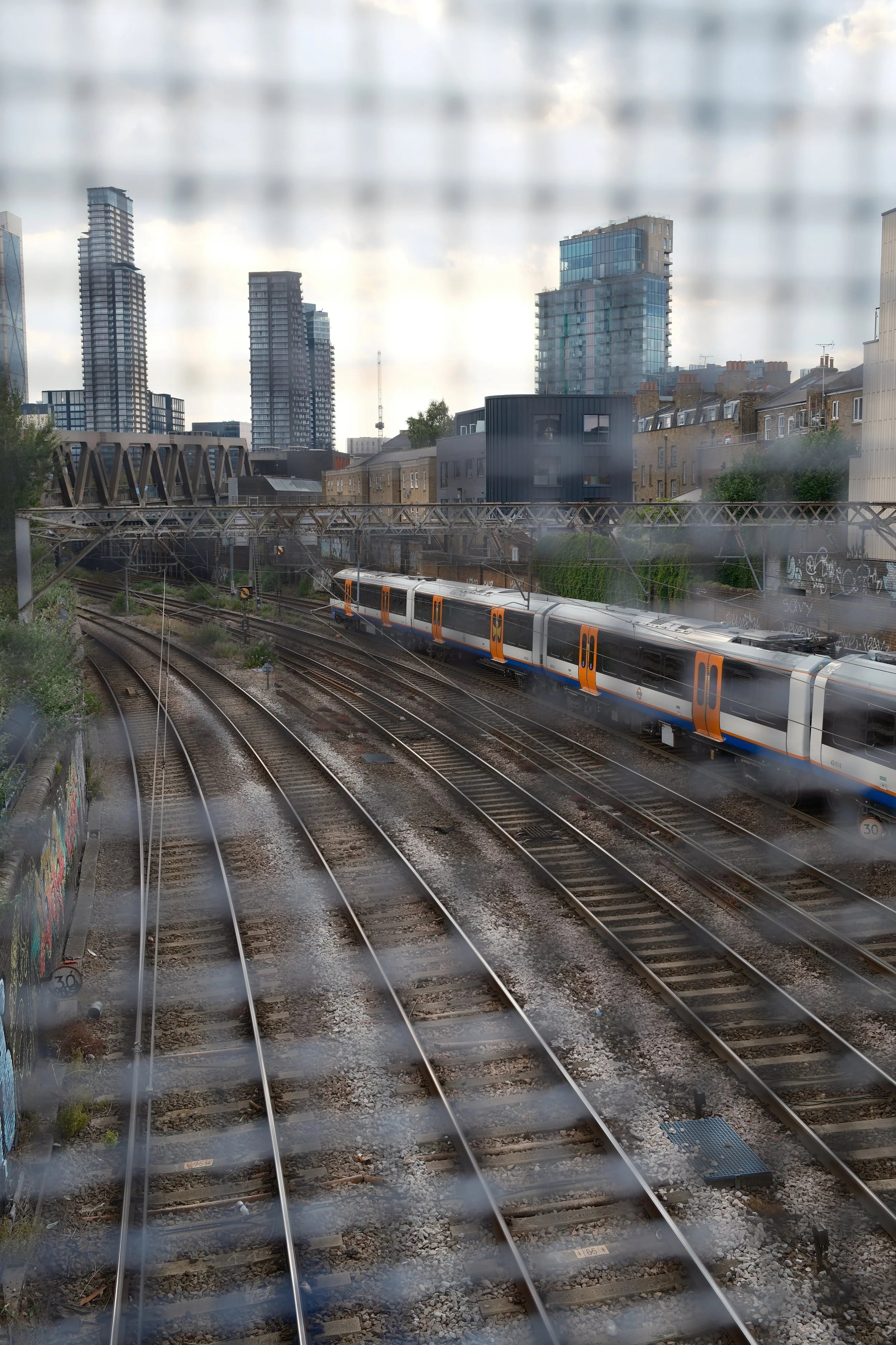 A view of train tracks with a passing train in the foreground, and city buildings including high-rise apartments and commercial structures in the background, seen through a rain-covered window.