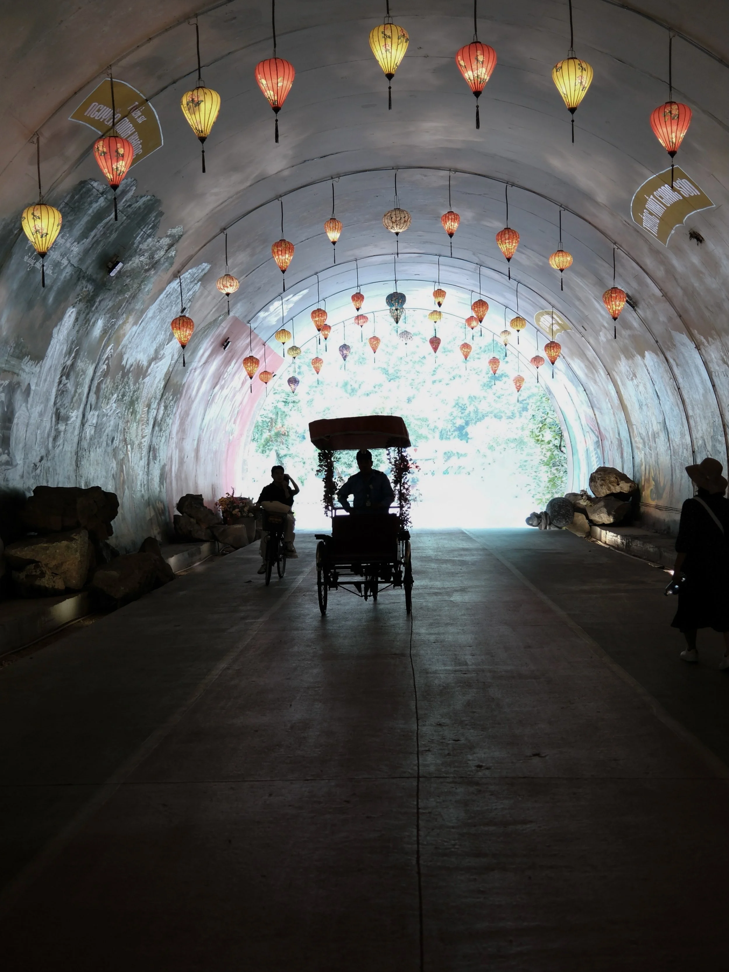 People walking and riding bikes inside a tunnel decorated with colorful hanging lanterns, with a bright opening at the end of the tunnel, and rocks placed on the ground.