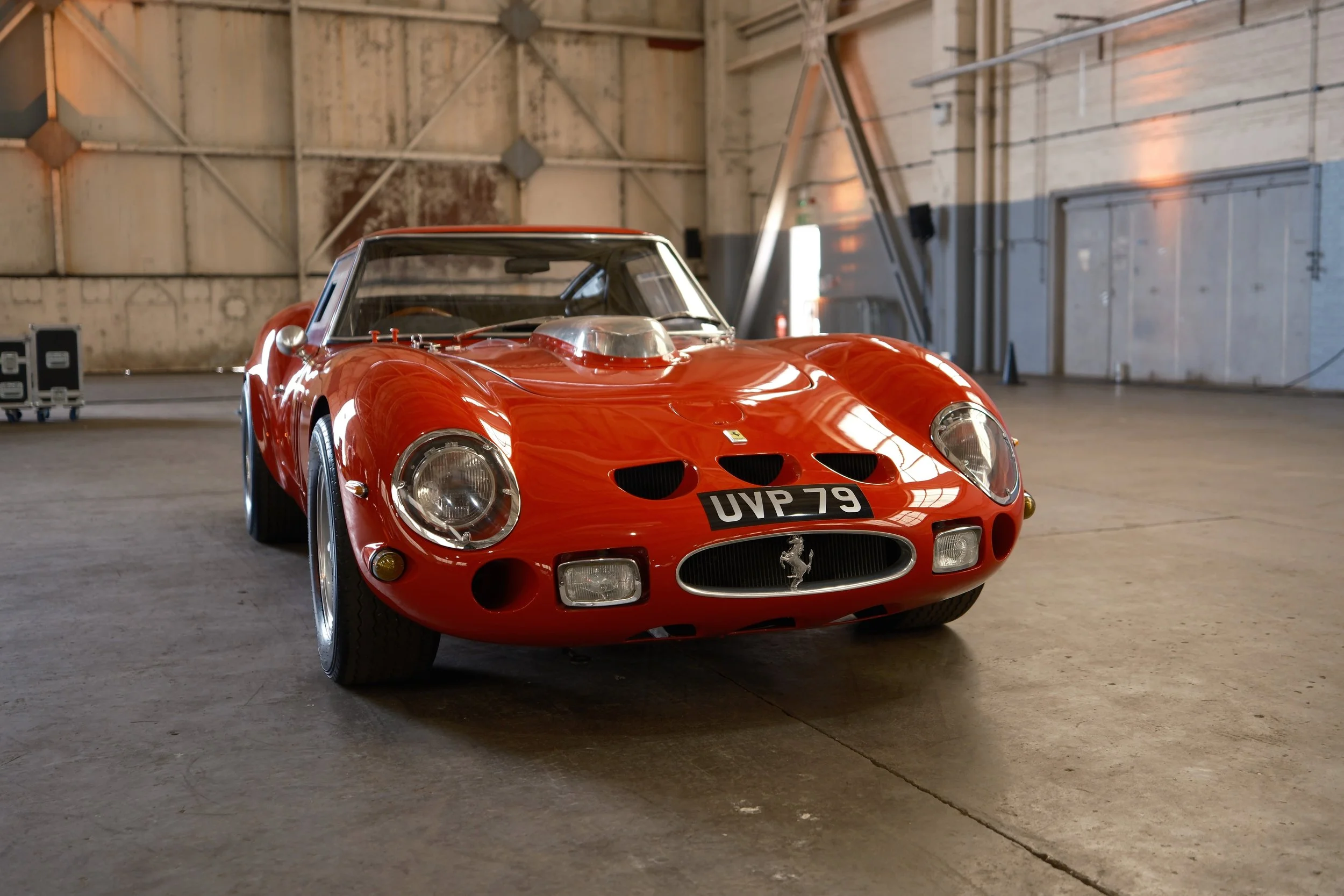 A red vintage Ferrari race car with a silver hood scoop and black racing stripes, parked inside a large industrial warehouse with concrete floor and exposed wooden walls.