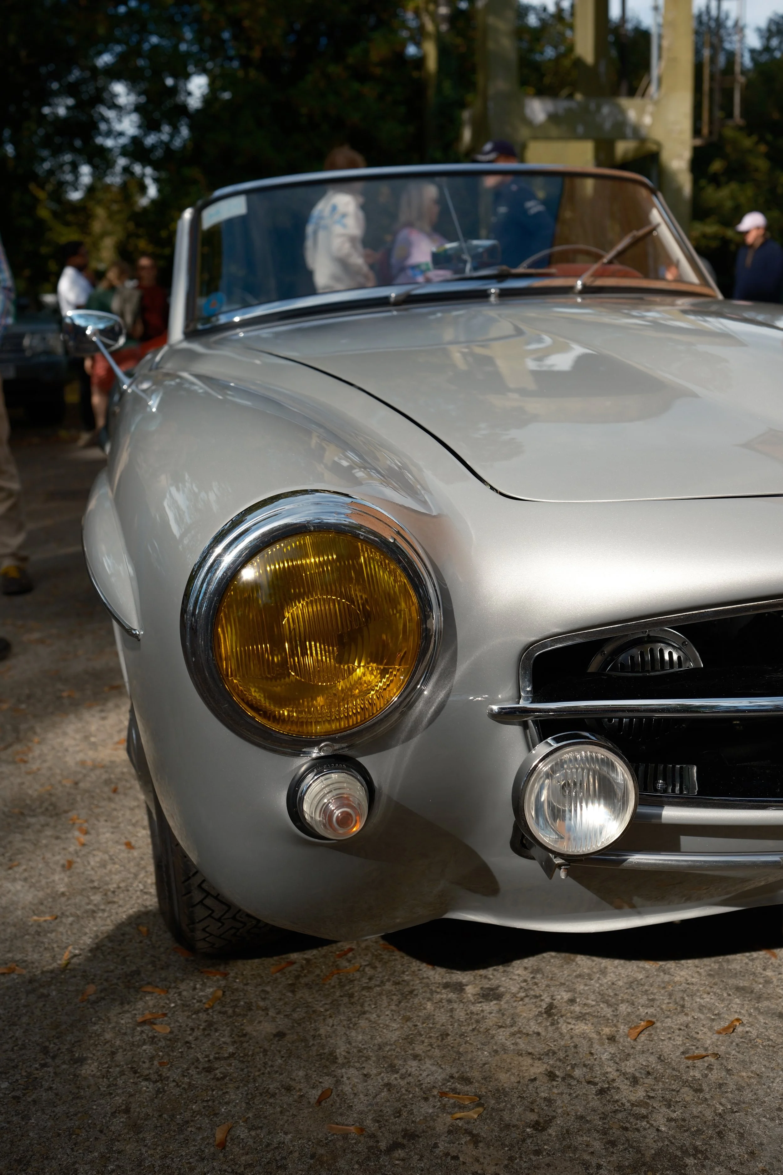 Close-up of the front of a silver vintage car with yellow-tinted headlights, parked outdoors on a gravel surface with fallen leaves.